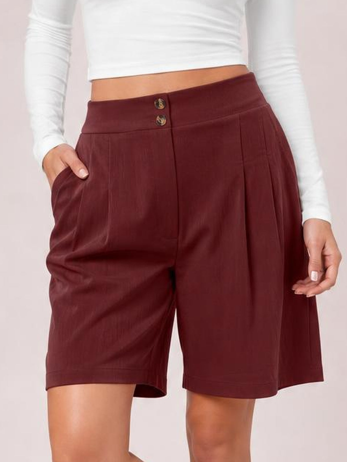 Model wears burgundy pocketed shorts with a wide-leg silhouette, knee-length hems, front pleats, and a buttoned waistband, one hand tucked into a pocket.