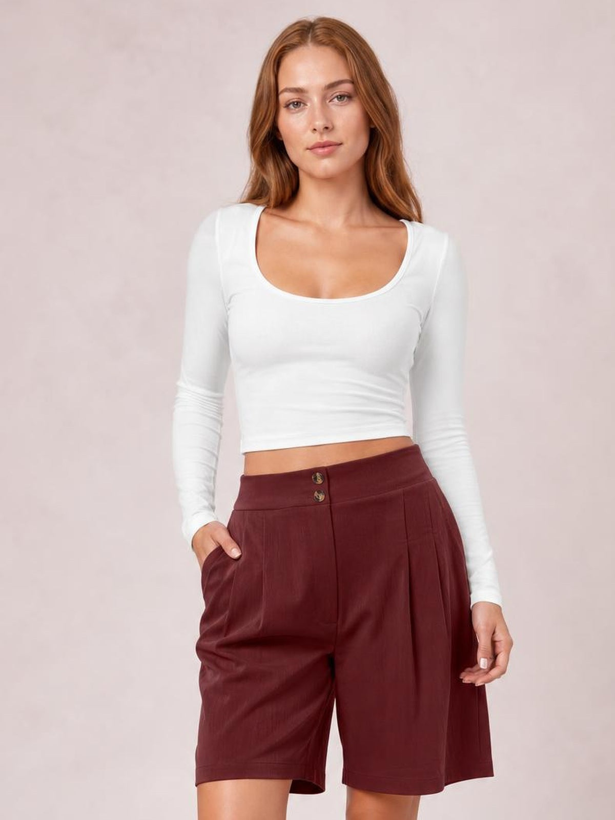 A woman modeling a white long-sleeve scoop-neck crop top and high-waisted maroon shorts against a soft pink studio background.