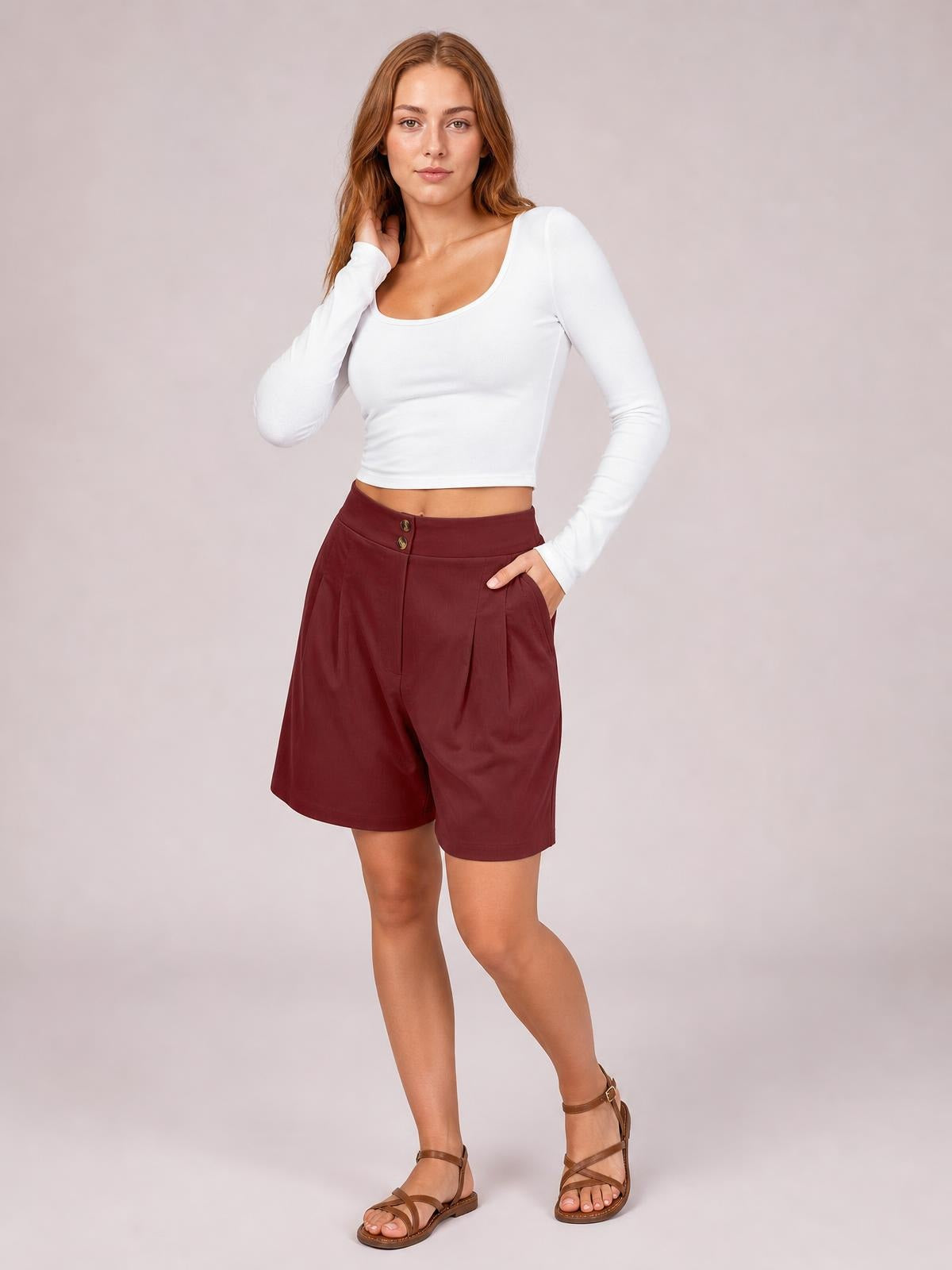 A model poses in burgundy pocketed shorts with a structured waistband, front buttons, soft pleats, and a relaxed knee-length silhouette.