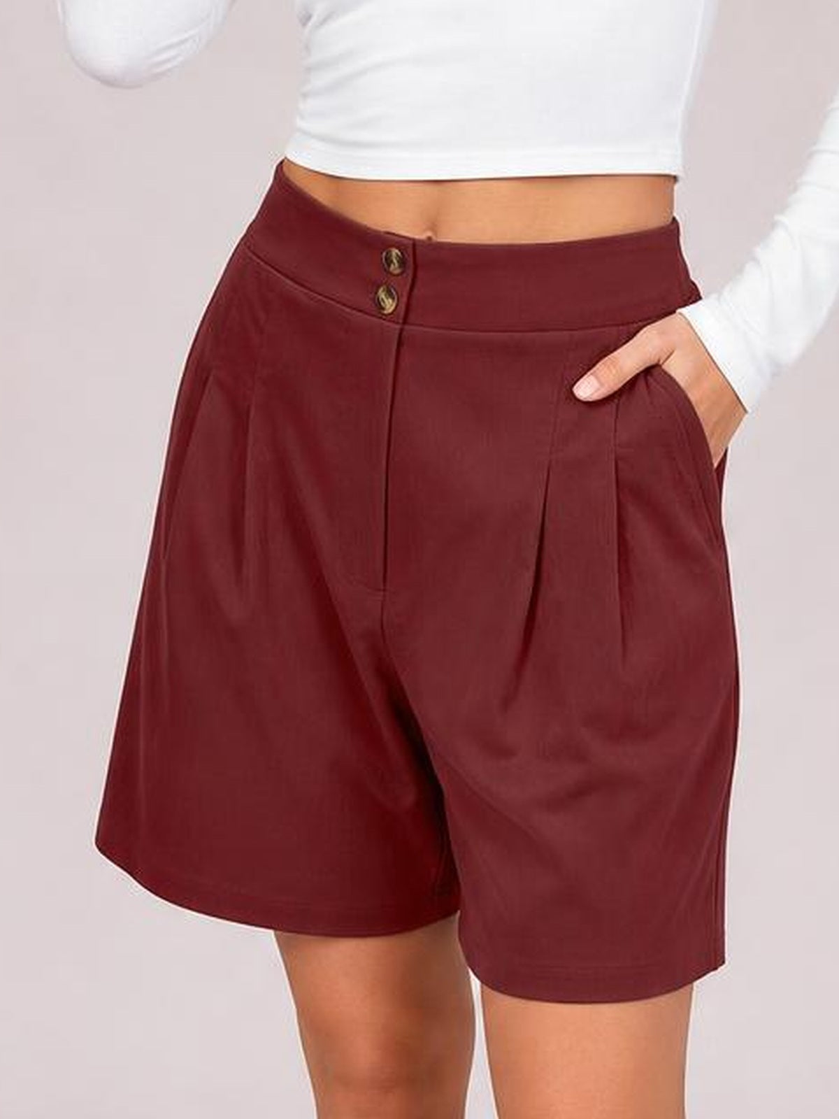 A model wears burgundy pocketed shorts with a relaxed wide-leg silhouette and two-button front closure, posing with one hand in a side pocket.