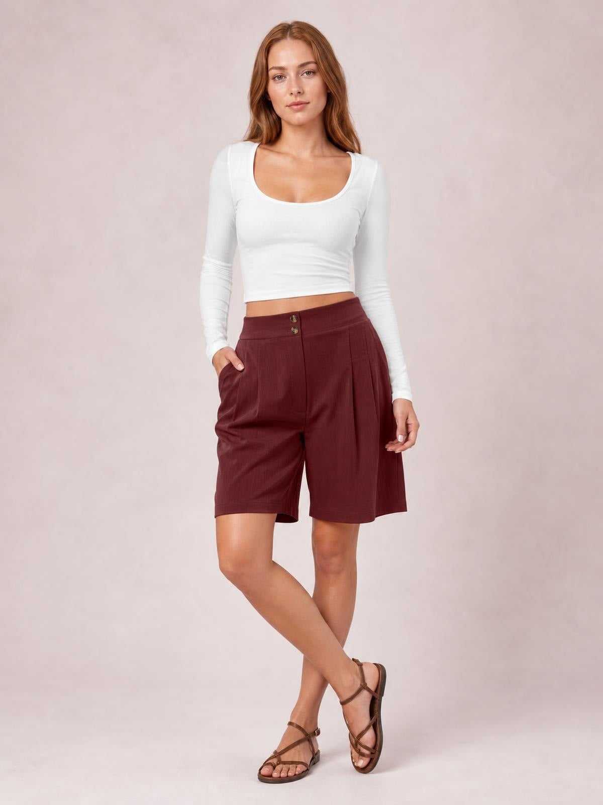 A model poses with crossed legs in burgundy pocketed shorts featuring a high waistband, button closure, subtle pleats, and a relaxed knee-length silhouette.