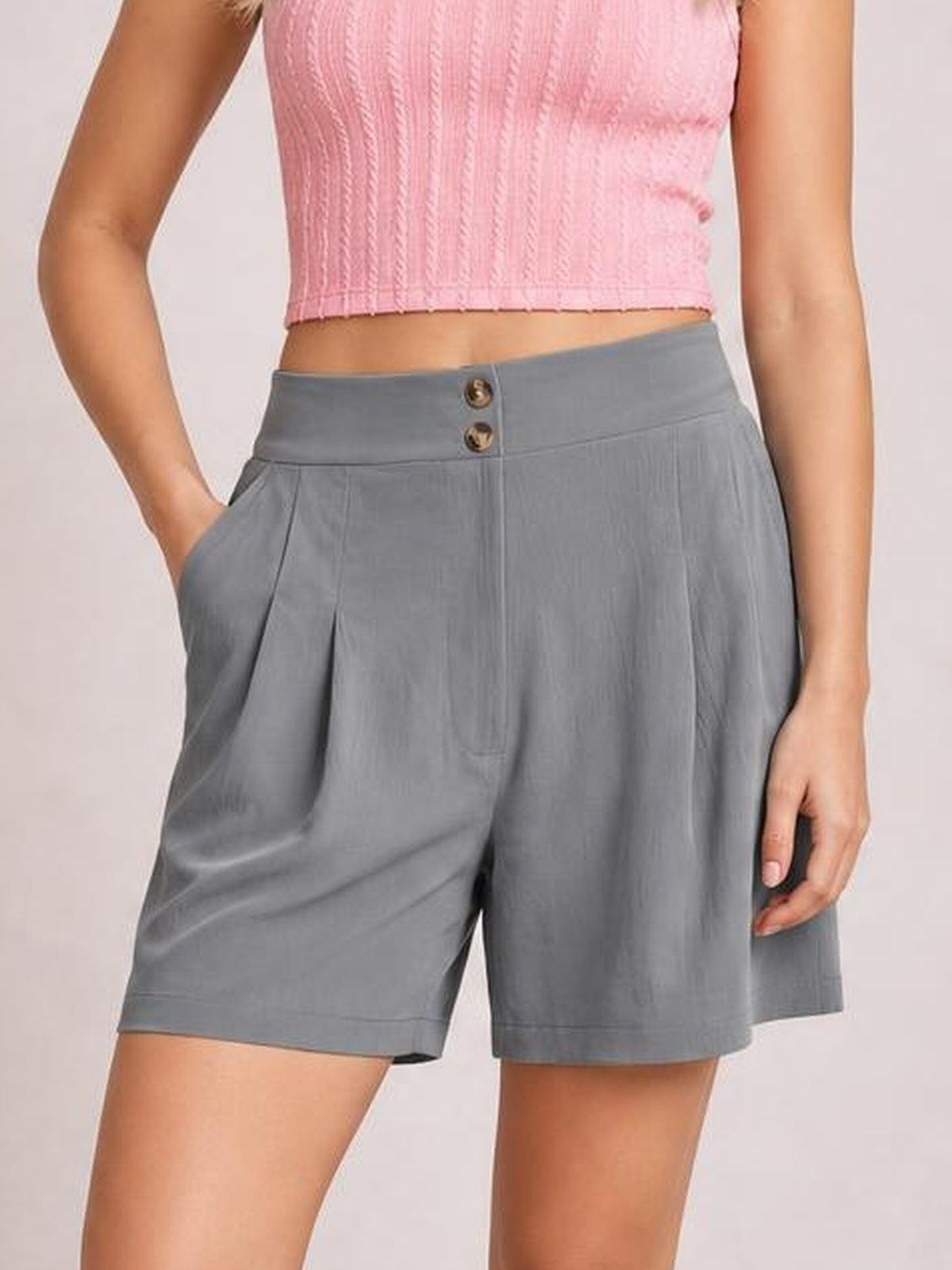 A model wears gray pocketed shorts with a relaxed wide-leg silhouette, two-button waistband, front pleats, and knee-length construction while posing with one hand in a pocket.