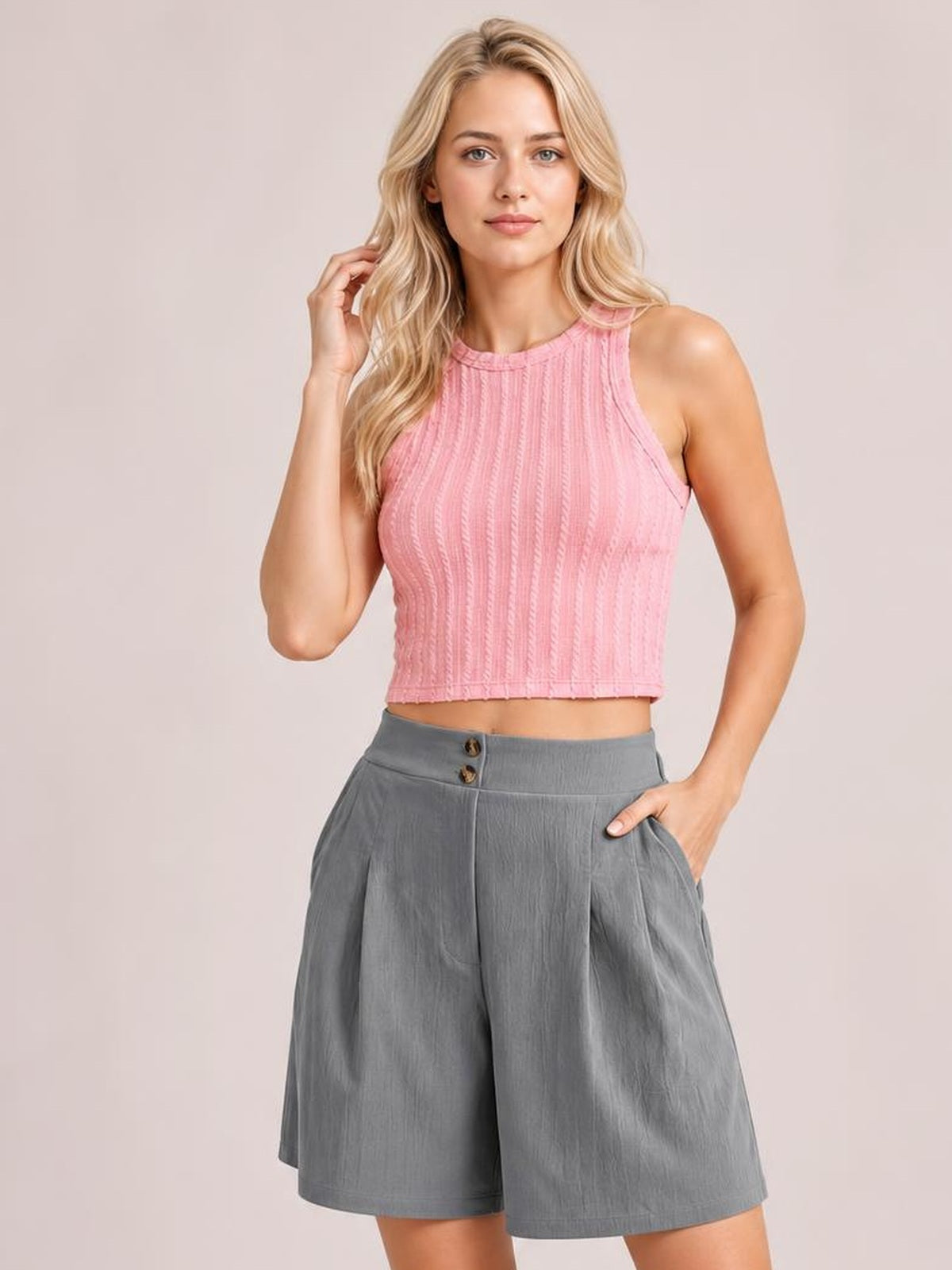 A blonde model wearing a pink ribbed knit sleeveless crop top and gray pleated shorts, standing against a solid light beige studio background.