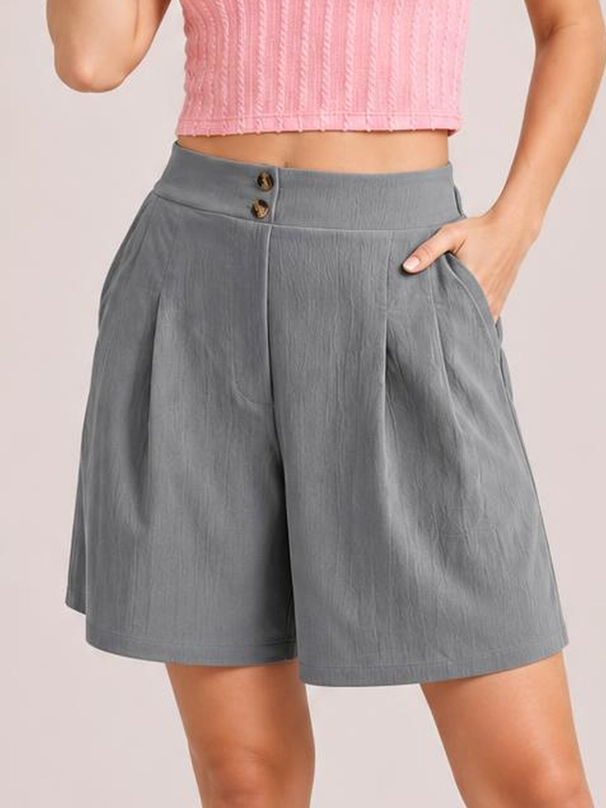 A model wears gray pocketed shorts with a structured waistband, two front buttons, relaxed wide legs, and one hand tucked into a side pocket.