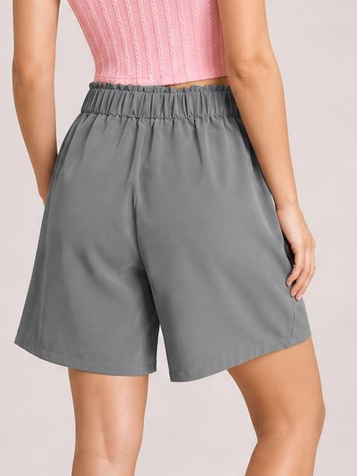 A model wears gray pocketed shorts with a ruched half-elastic waistband, relaxed wide-leg silhouette, smooth opaque fabric, and mid-thigh hem.