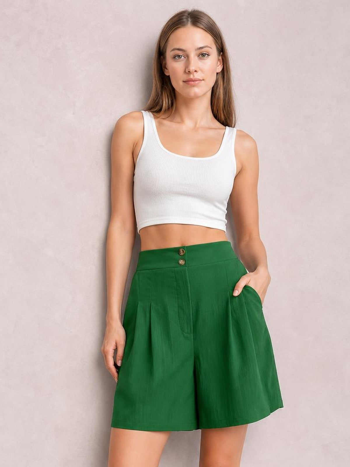 A woman modeling a white ribbed crop tank top and high-waisted green shorts against a textured light gray studio backdrop.