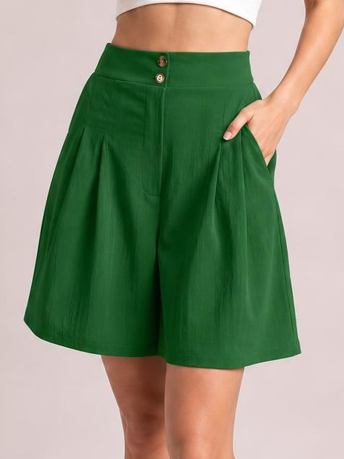 A model wears deep green high-waisted shorts with a wide-leg silhouette, buttoned waistband, side pockets, and a softly pleated construction.