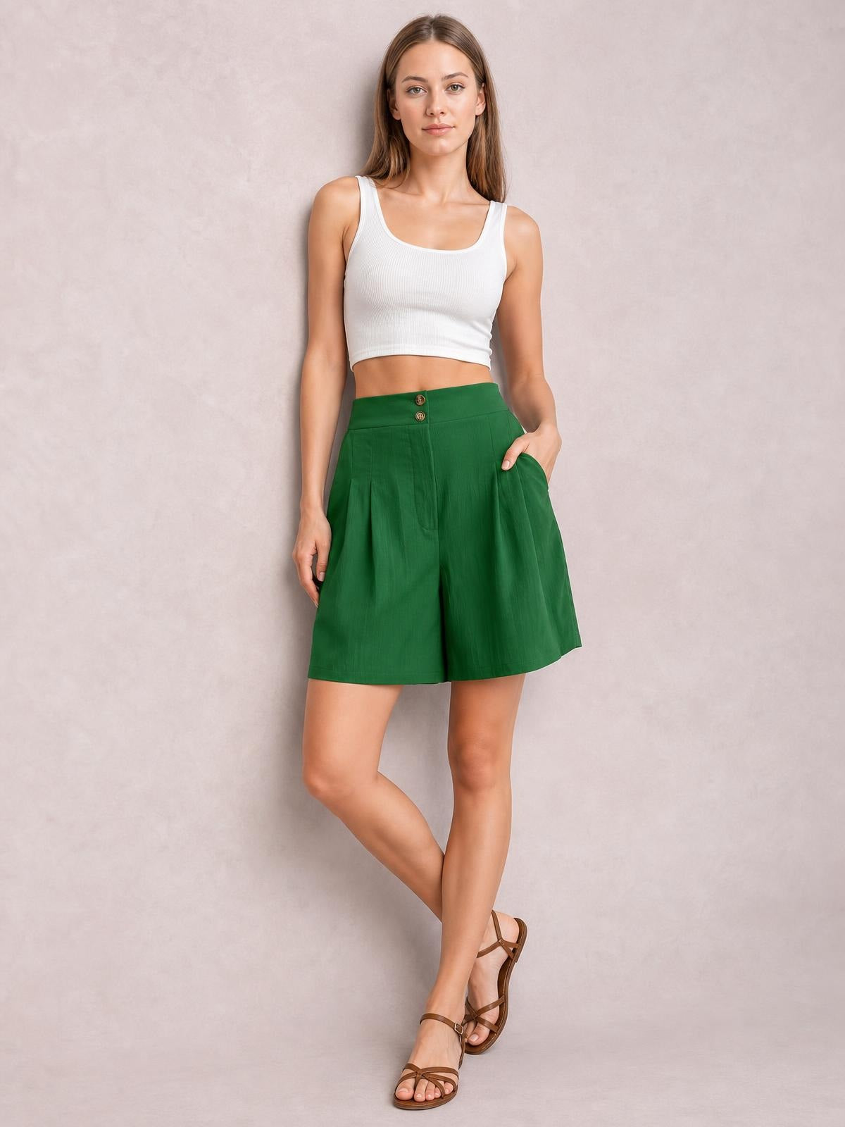 A model wears green pocketed shorts with a high half-elastic waist, button front, pleated construction, and a relaxed wide-leg silhouette while posing with crossed legs.