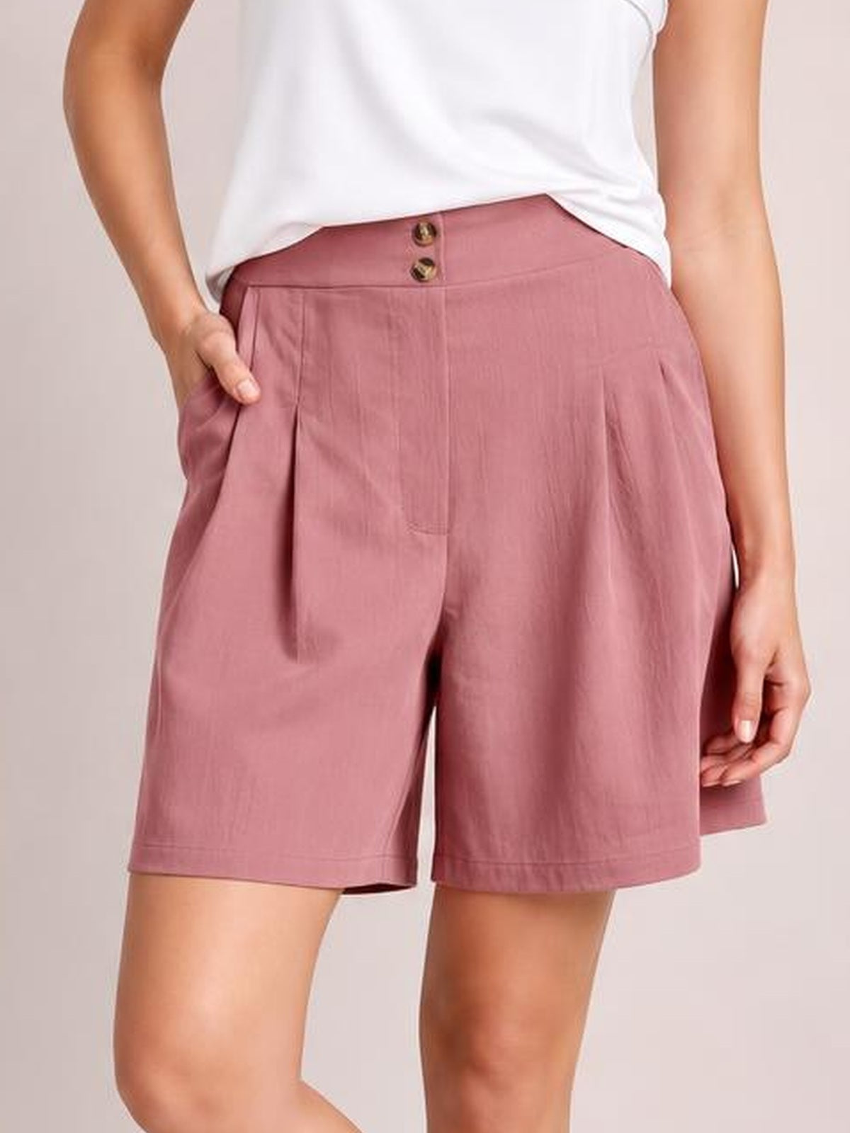 A model poses in dusty rose pocketed shorts with a relaxed wide-leg silhouette, front pleats, buttoned waistband, and one hand tucked into a pocket.