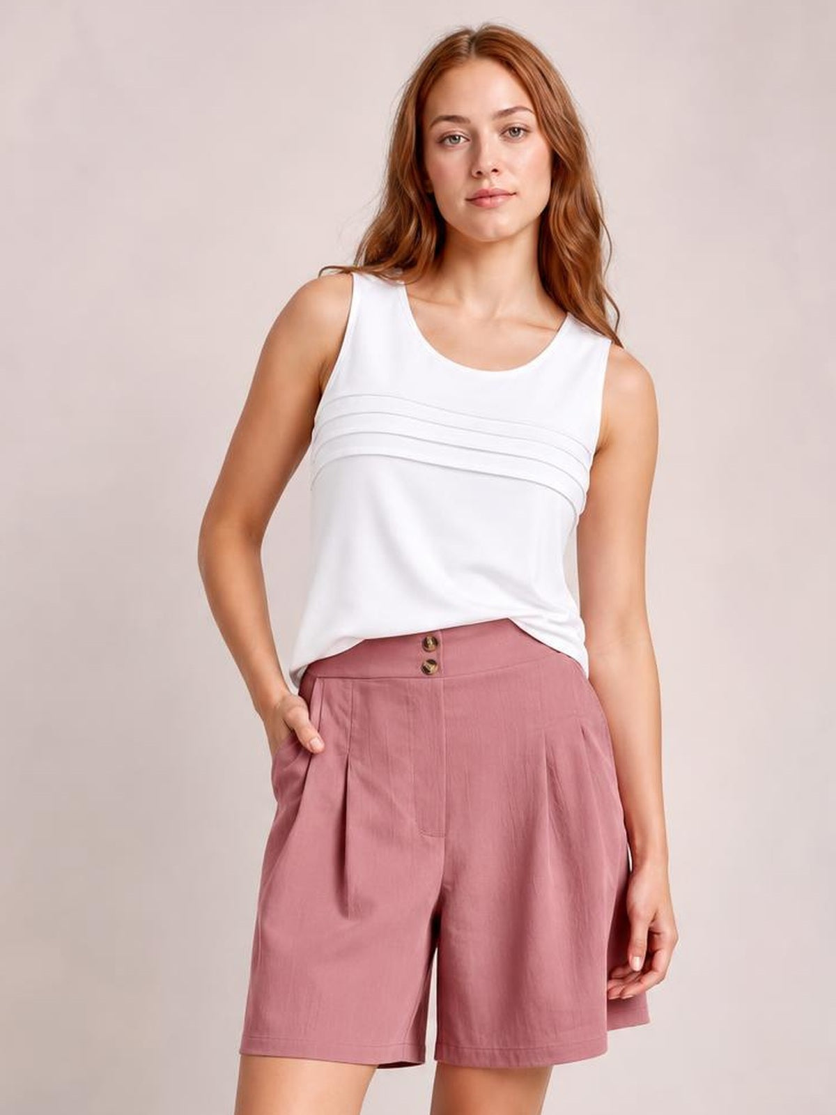 A woman modeling a white sleeveless top with horizontal pleats and dusty rose pleated shorts against a plain beige studio background.