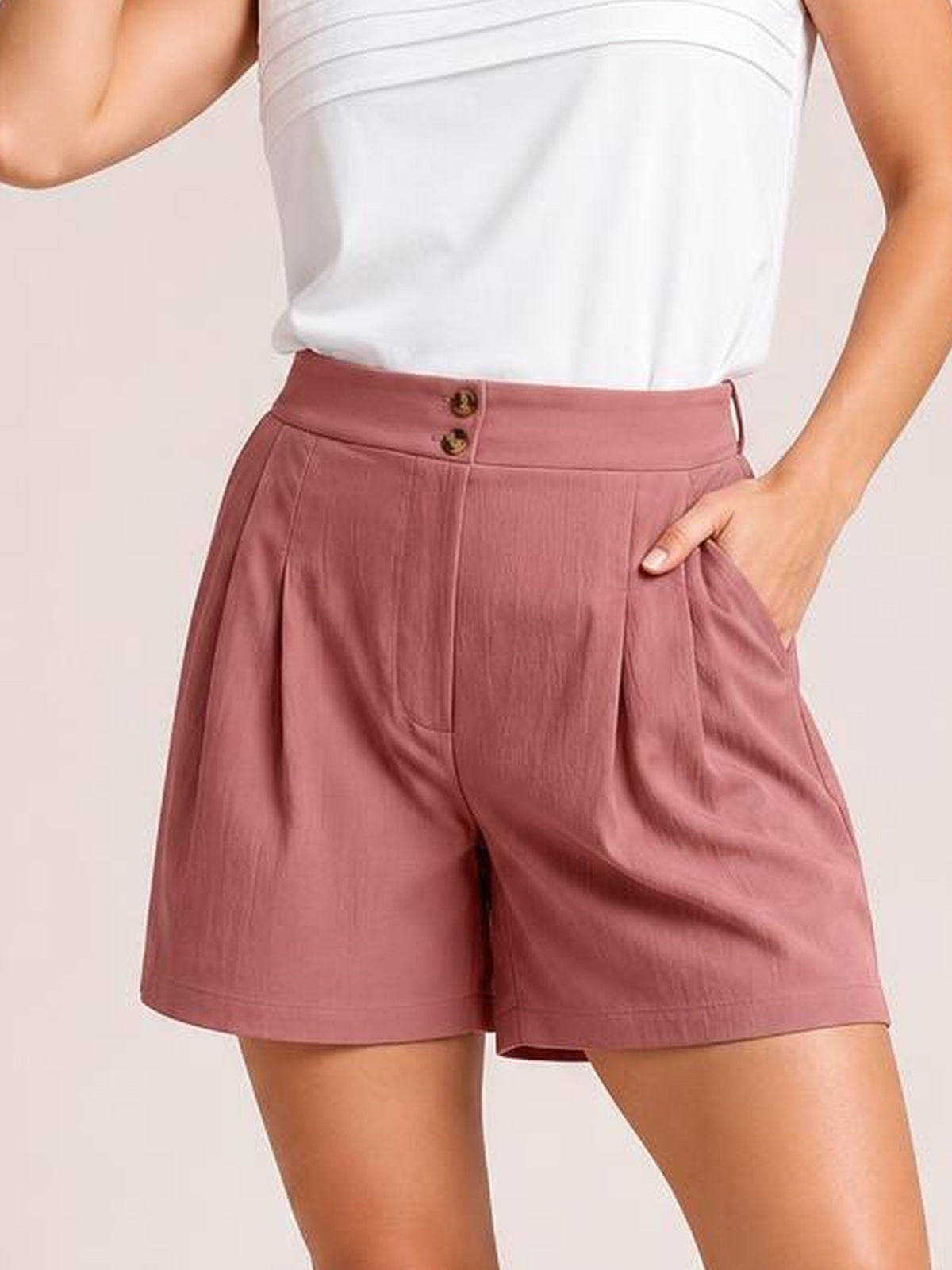 A model wears dusty rose pocketed shorts with a relaxed wide-leg silhouette, buttoned waistband, front pleats, and one hand tucked into a pocket.