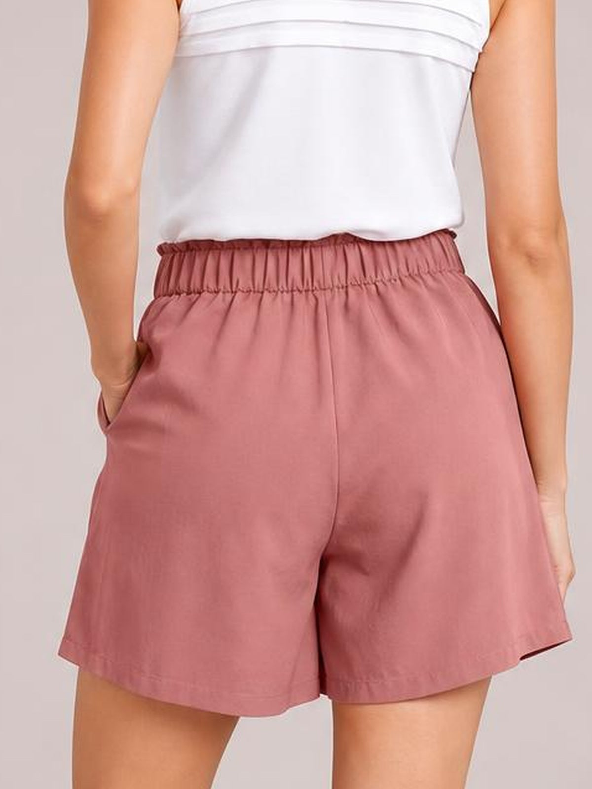 A model wears dusty rose pocketed shorts with a ruched elastic waistband, relaxed loose silhouette, and straight hems, shown from the back with one hand in a pocket.