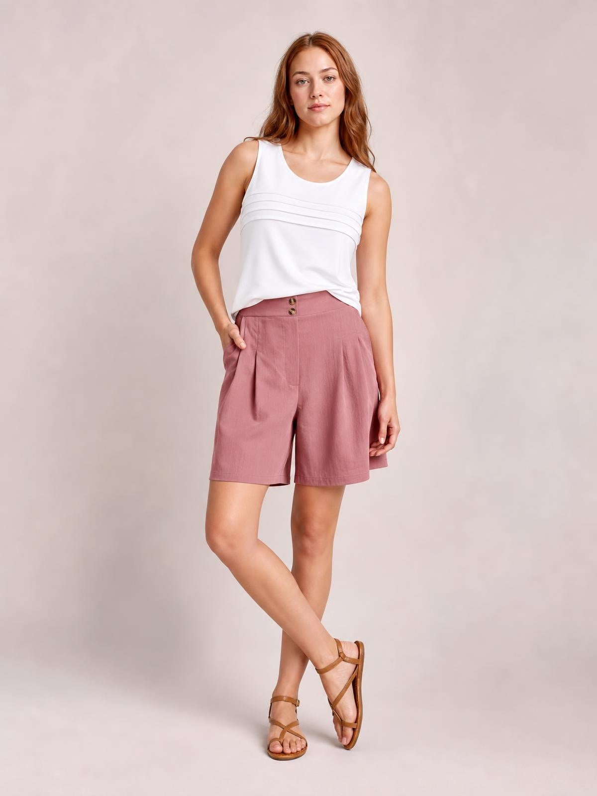 A model poses in dusty rose pocketed shorts with a straight knee-length silhouette, front pleats, and a buttoned waistband, one hand resting in a pocket.
