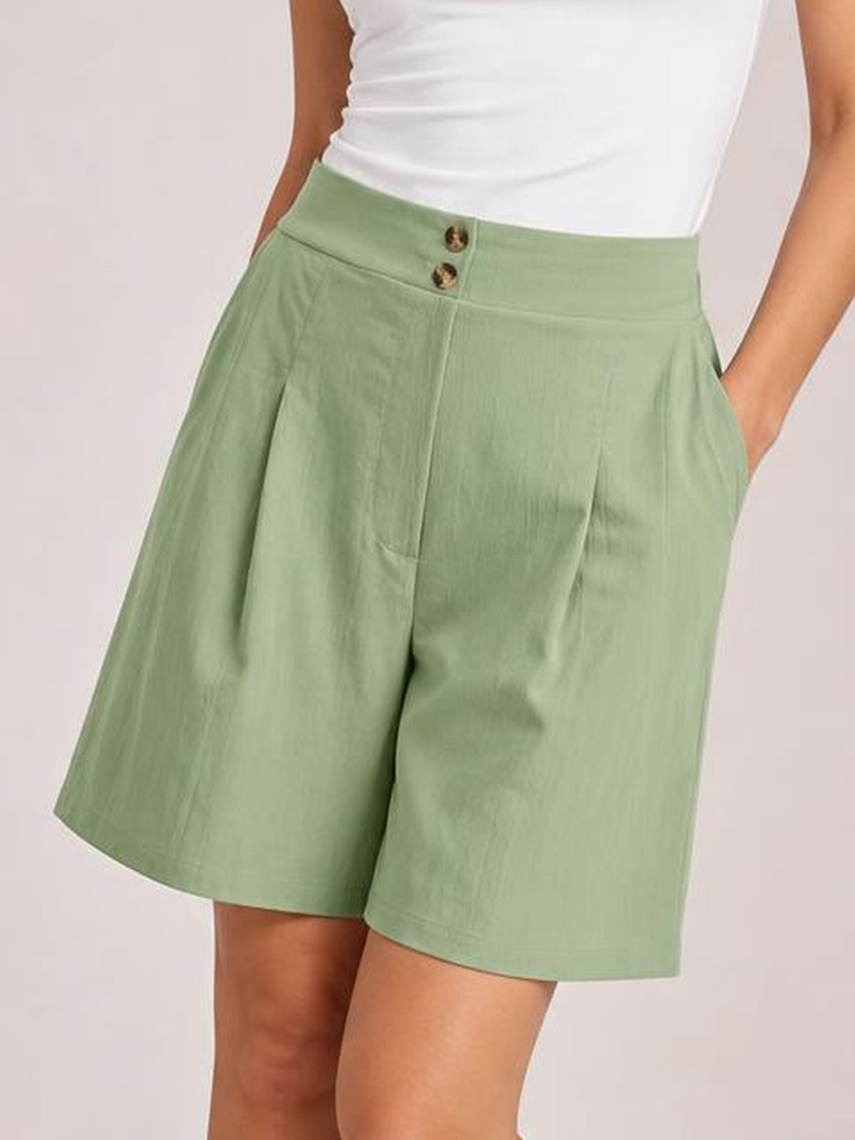 A model poses with one hand in a pocket, showing sage green pocketed shorts with a high waistband, button front, wide-leg silhouette, and knee-length hem.