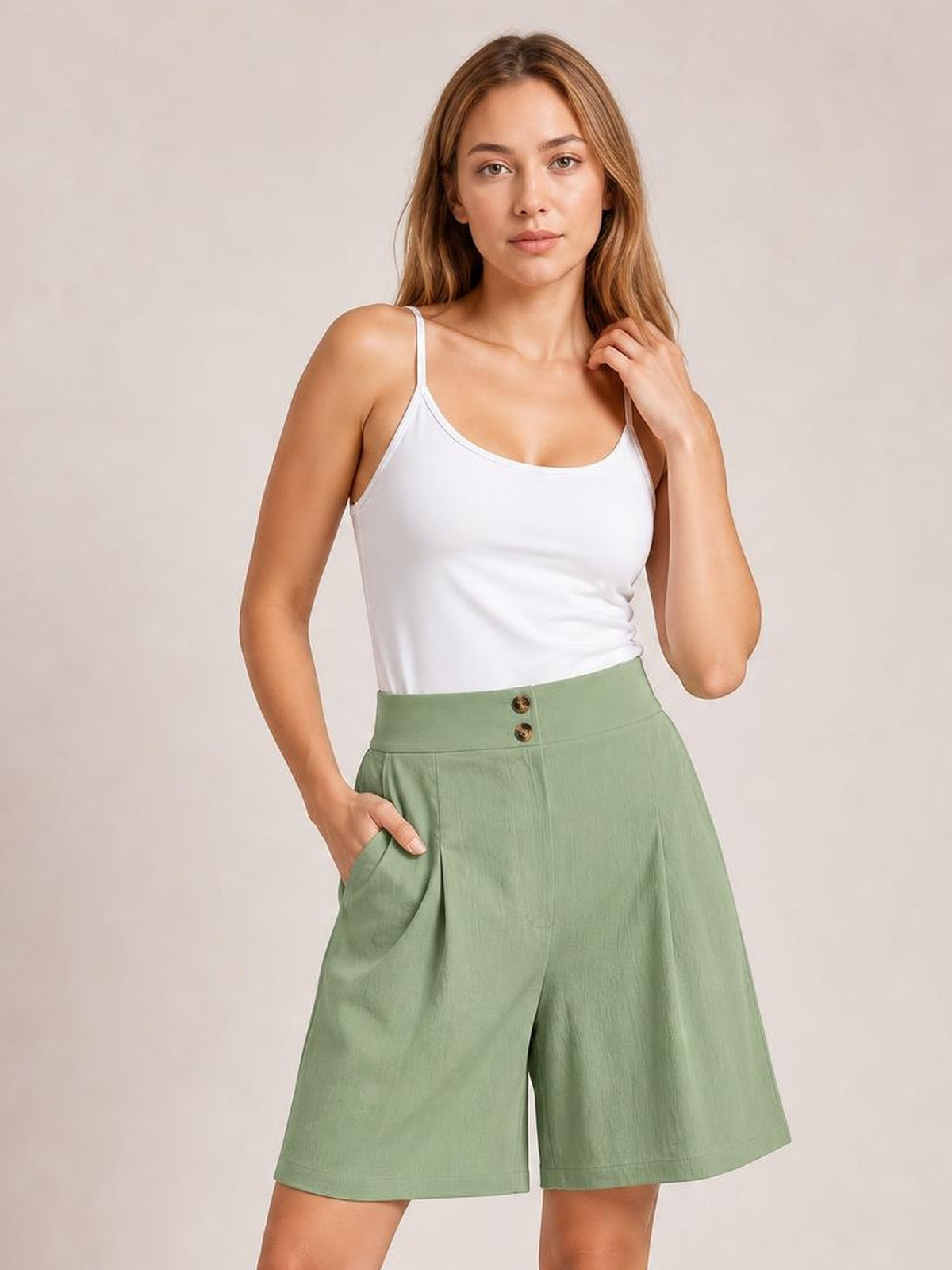 A woman modeling sage green high-waisted shorts with a white camisole top, standing against a neutral beige studio background.