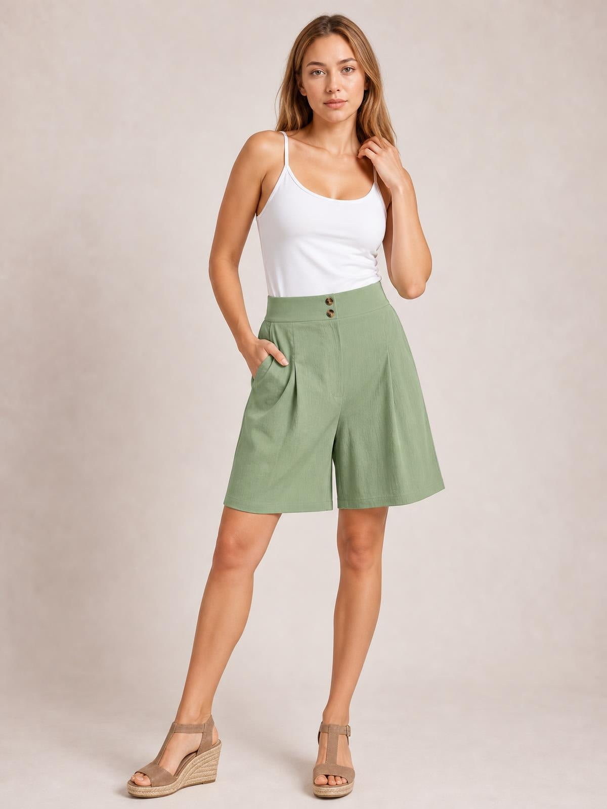 A model poses in sage green pocketed shorts with a high waist, front button closure, wide-leg silhouette, and knee-length hem.
