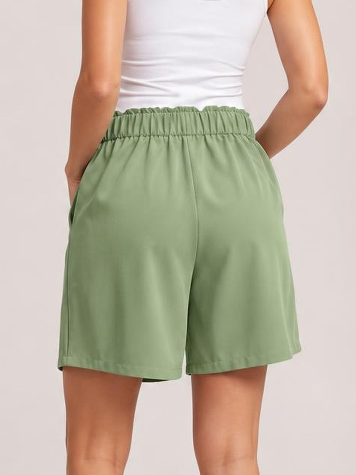A model poses with hands in the pockets, showing sage green shorts with a ruched half-elastic waistband and relaxed, straight-leg silhouette.