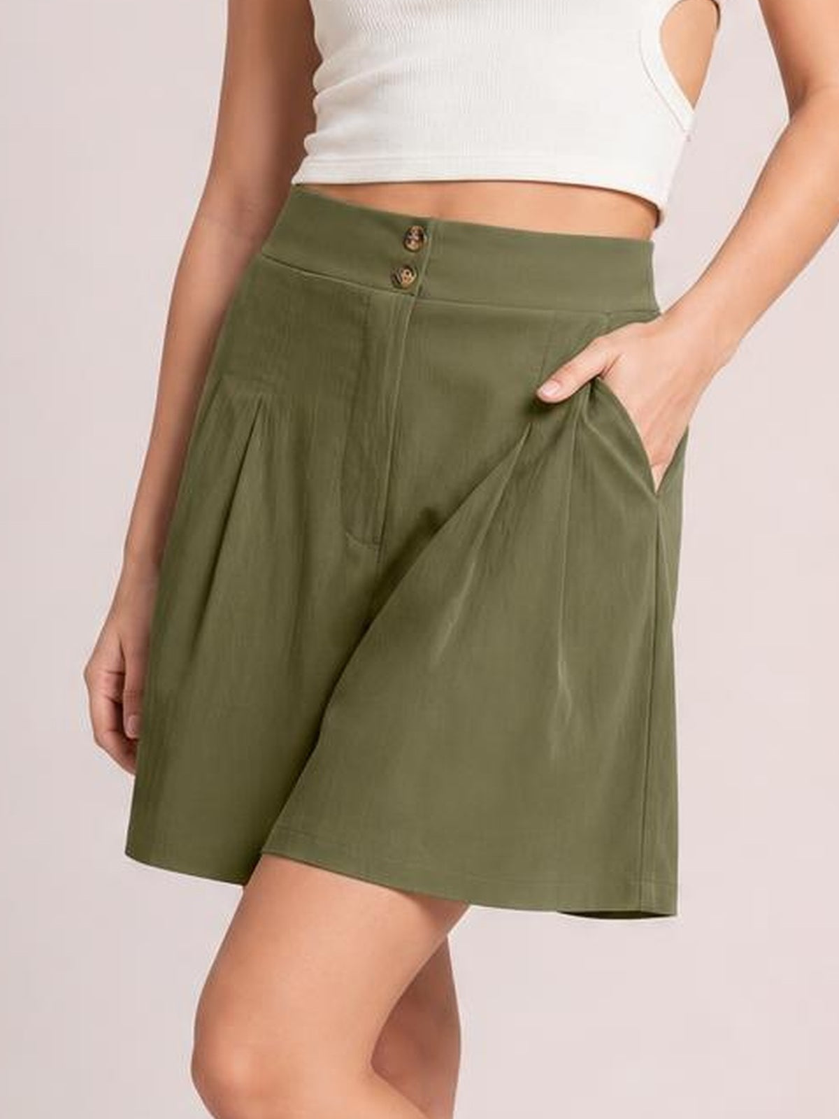 A model wears olive green pocketed shorts with a buttoned waistband, ruched front pleats, and a relaxed wide-leg silhouette while posing with one hand in a pocket.