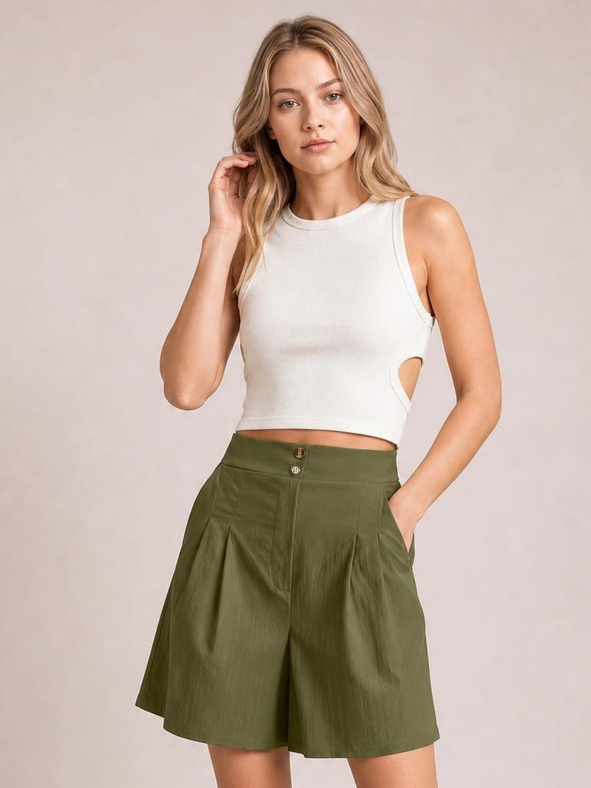 A woman modeling a white ribbed sleeveless crop top with side cutouts and olive green high-waisted pleated shorts against a solid light beige studio background.