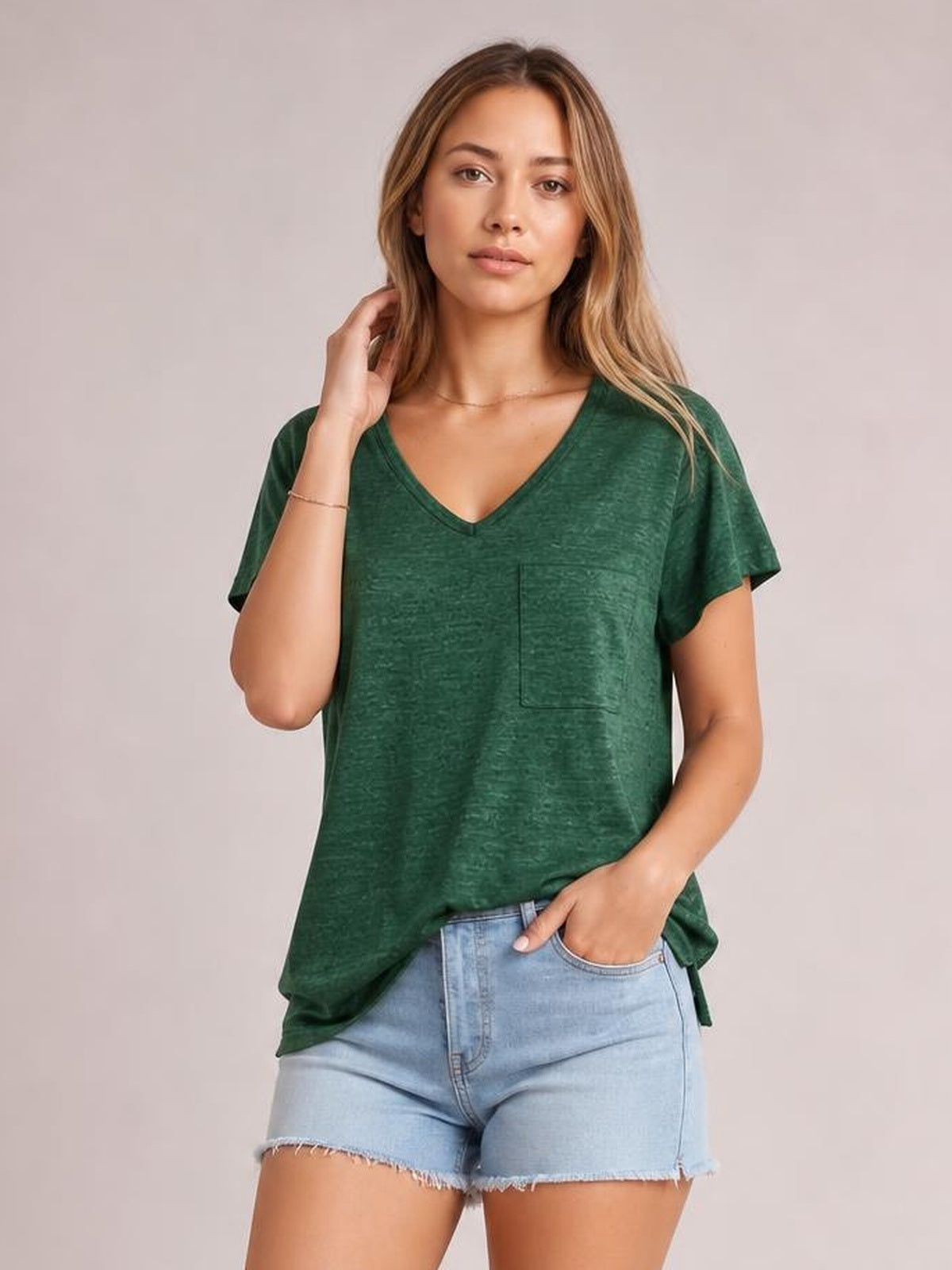 A model poses in a relaxed, heathered green V-neck short-sleeve T-shirt with a loose silhouette and visible front patch pocket.