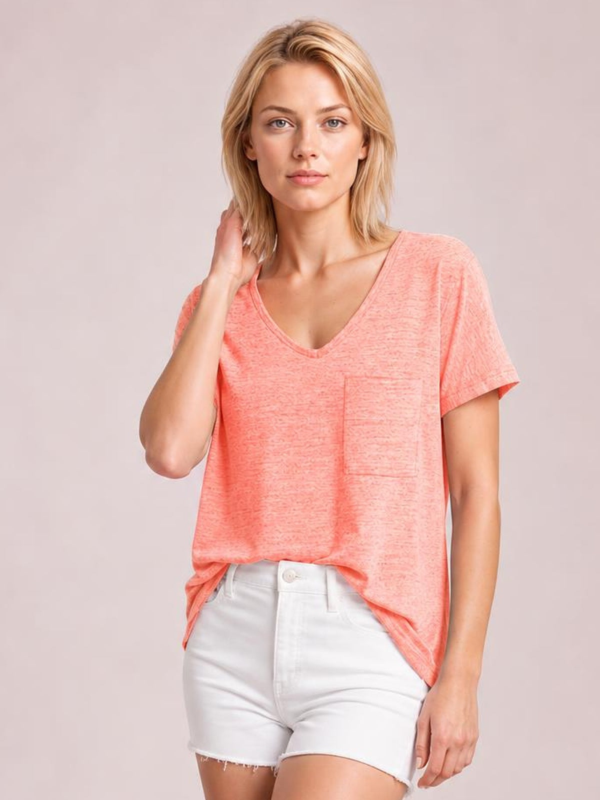 Model poses in a coral pink heathered V-neck short sleeve T-shirt with a chest pocket and relaxed silhouette, lightly tucked into white shorts.