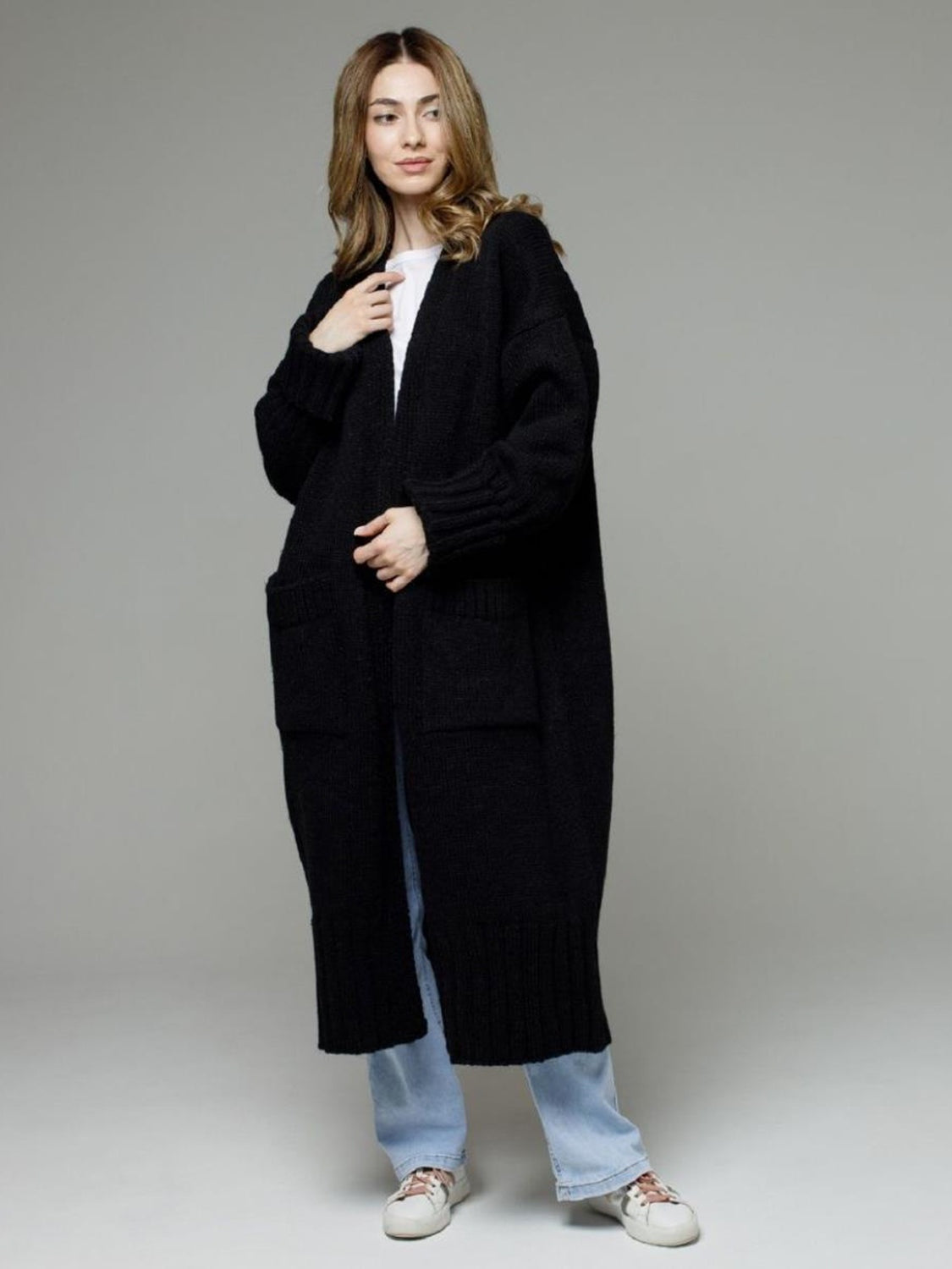 Model poses in a long black pocketed open-front cardigan with dropped shoulders, oversized sleeves, ribbed cuffs, and a relaxed silhouette over jeans.