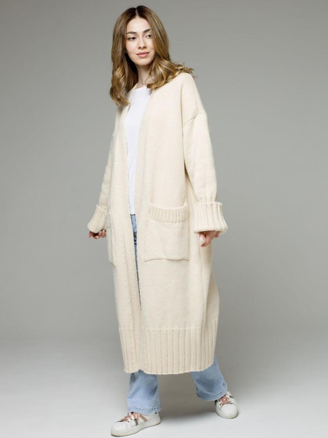 A model stands in a cream open-front cardigan with dropped shoulders, long relaxed silhouette, ribbed cuffs and hem, and large front pockets.