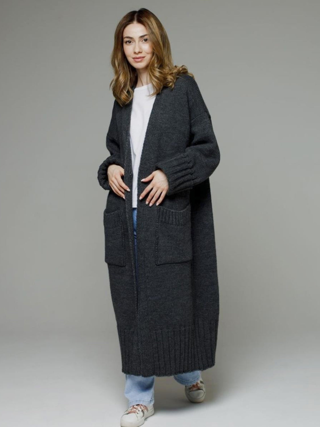 A model stands in a dark gray pocketed open-front cardigan with dropped shoulders, long relaxed silhouette, ribbed cuffs and hem, and oversized patch pockets.