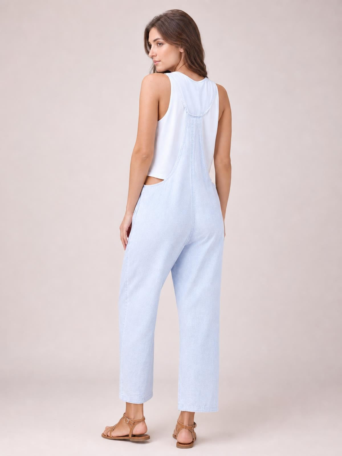 Model shown from behind wearing light blue denim overalls with a low back bib, wide cropped legs, curved seams, and a relaxed straight silhouette.