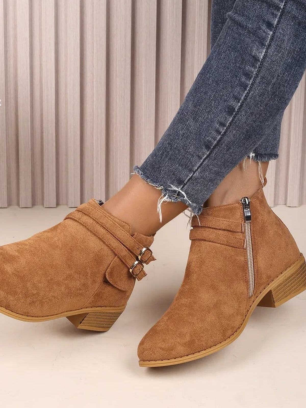 Tan suede point-toe block heel boots with ankle height, side zippers, double buckle straps, and textured rubber soles shown with cropped jeans.