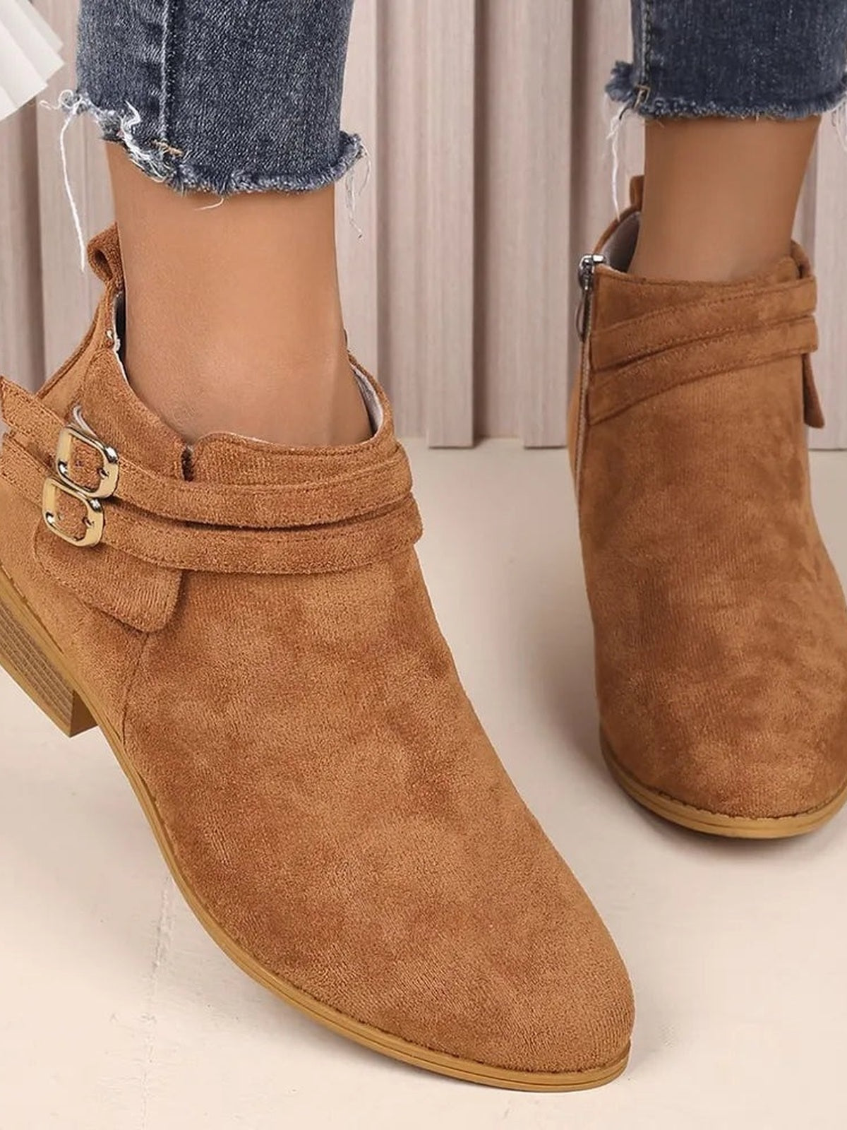 A model wears tan suede point-toe ankle boots with low block heels, wraparound straps, and gold-tone buckles over cropped frayed jeans.