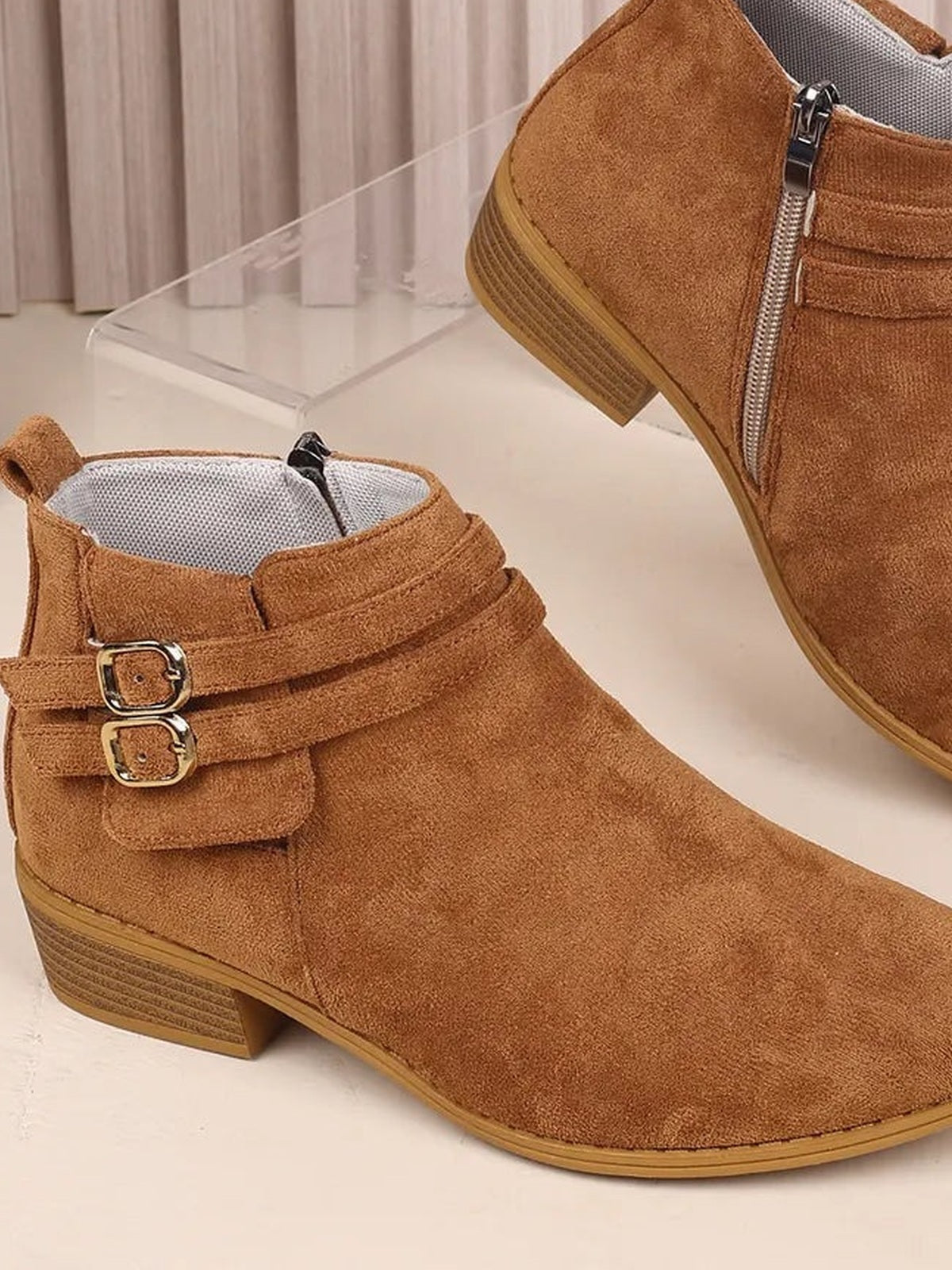 Brown suede point-toe ankle boots feature low block heels, double buckle straps, side zippers, and textured rubber soles.