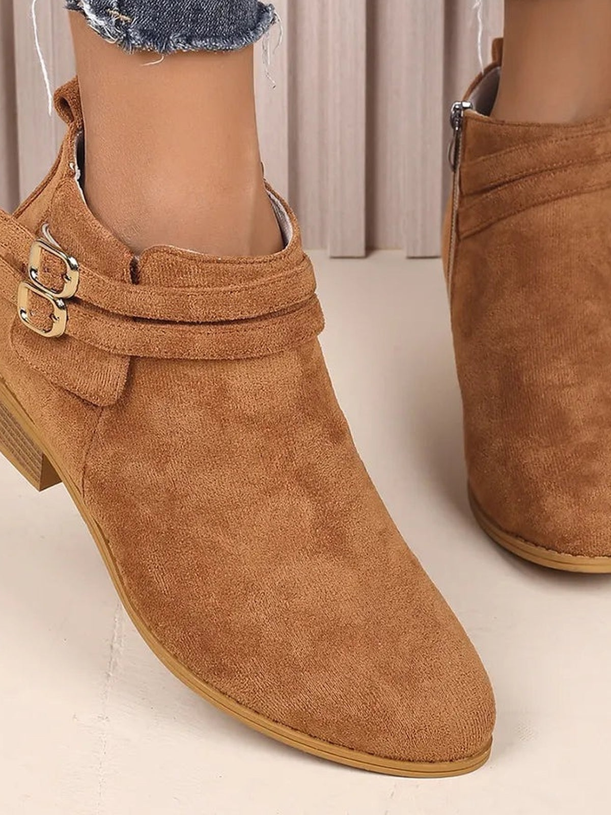 Close-up of brown suede point-toe boots on a model, featuring low block heels, double wraparound straps with buckles, and side zippers.