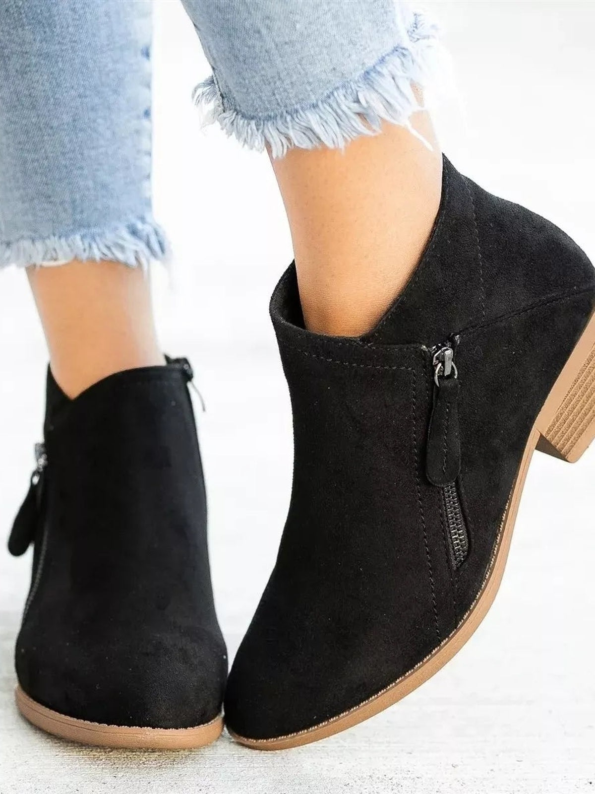 A model wears black ankle boots with side zippers, rounded toes, sturdy block heels, and tan rubber soles with cropped frayed jeans.