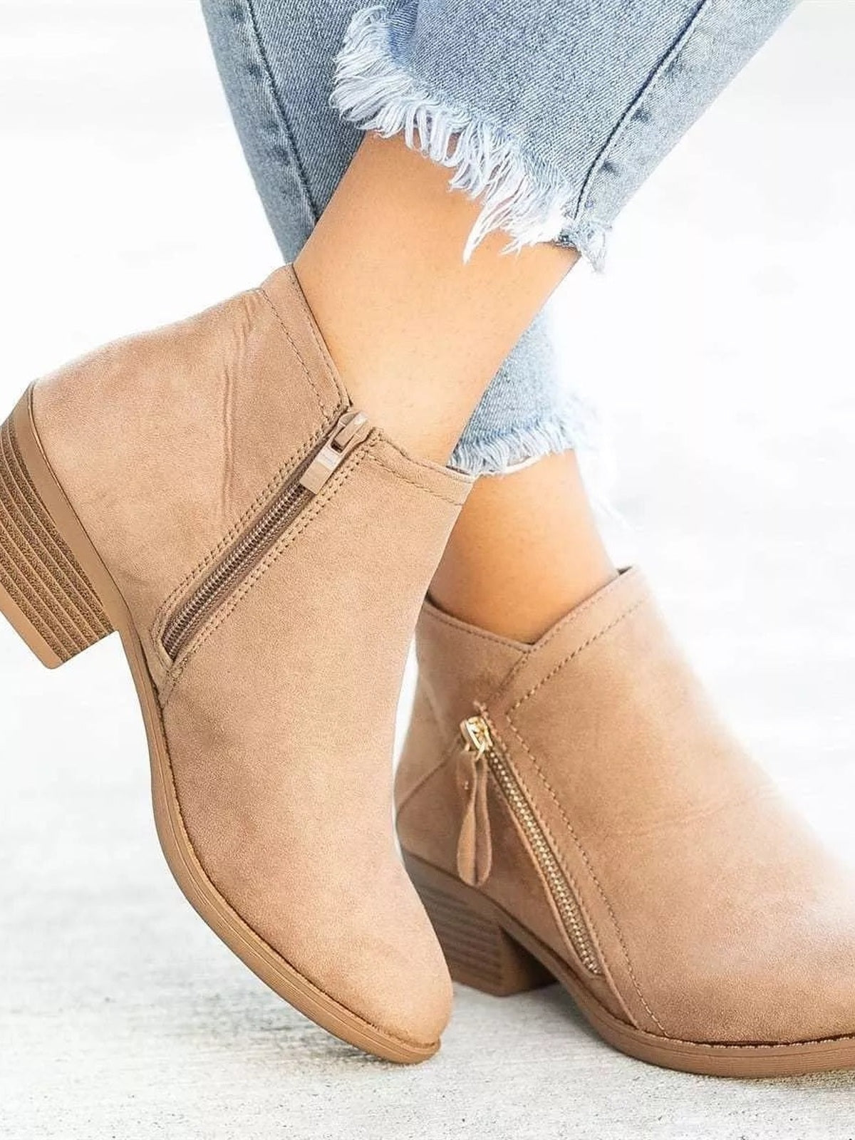 A model wears tan point-toe ankle boots with low block heels, side zippers, and stitched construction, posed with one foot crossed in front.