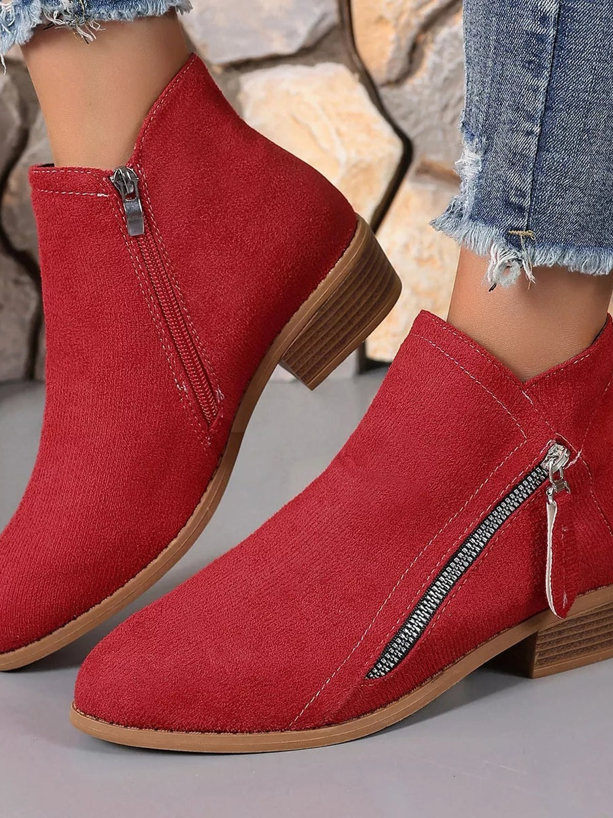 A model wears red point-toe ankle boots with side zippers, contrast stitching, and low stacked block heels beneath frayed cropped jeans.