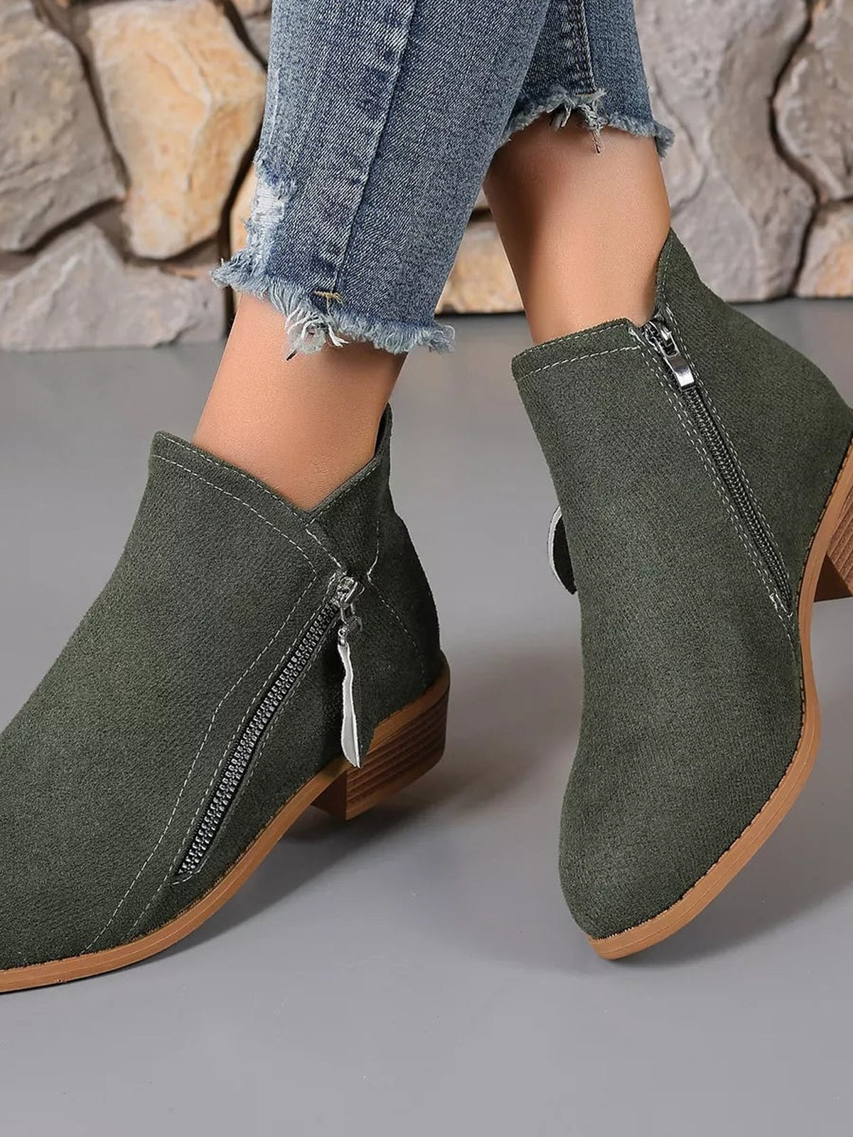 A model crosses her feet wearing olive-green point-toe ankle boots with low block heels, contrast stitching, side zippers, and tan rubber soles.