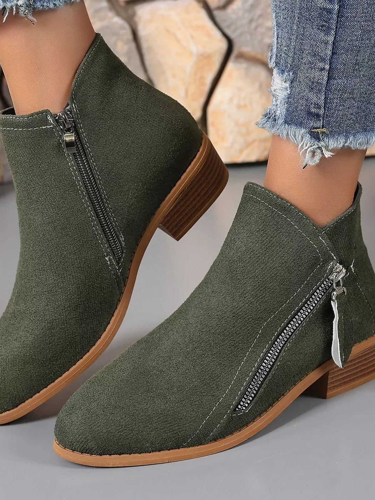 Olive-green point-toe ankle boots with low block heels, side zippers, contrast stitching, and textured uppers are shown on a model’s feet.