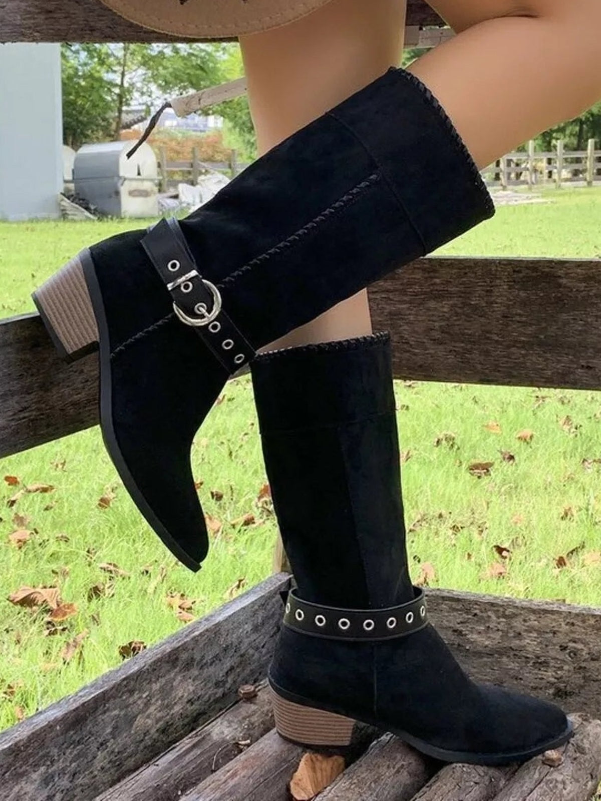A model wears black knee-high point-toe block heel boots with buckle straps, silver eyelets, stitched panels, and stacked mid heels, posing with one leg raised.