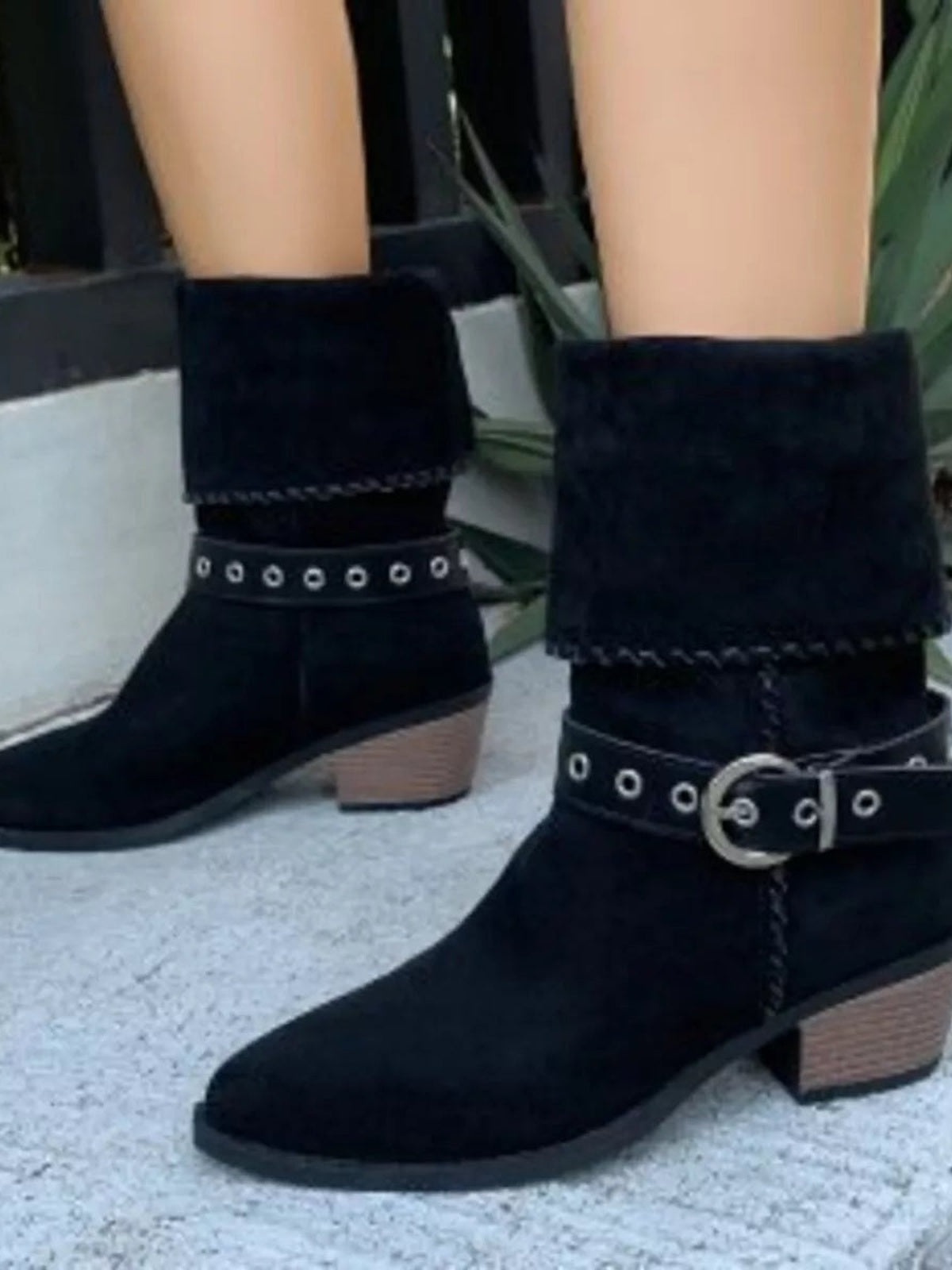 A model wears black ankle boots with pointed toes, mid block heels, layered cuff panels, and decorative eyelet straps with buckle closures.