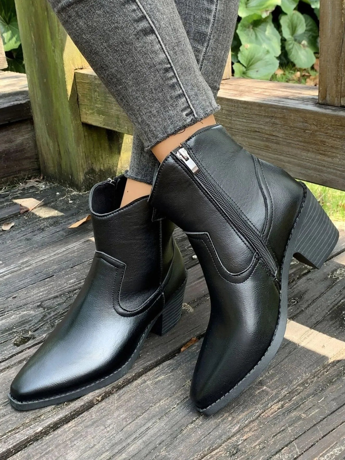 Black PU leather point-toe block-heel boots shown on crossed legs, featuring ankle height, side zippers, stitched panels, and sturdy rubber soles.