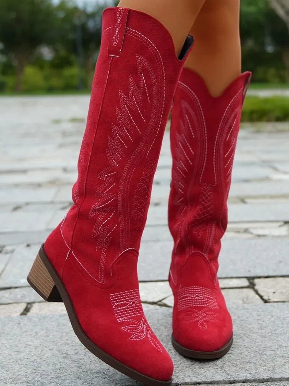 A model wears red suede point-toe boots with embroidered detailing, pull tabs, tall shafts, and mid-height block heels in a crossed-leg pose.