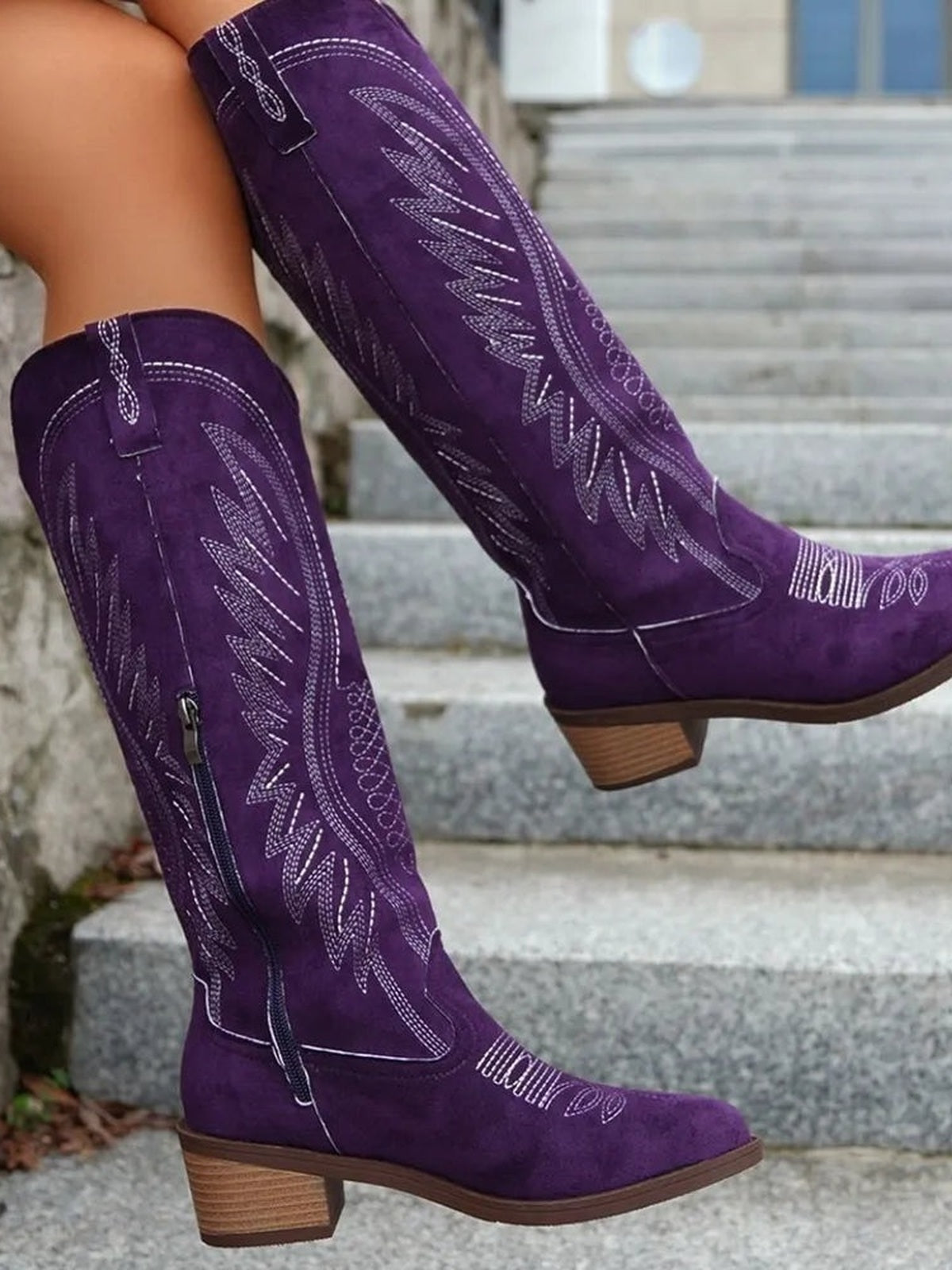 A model poses in tall purple suede boots with pointed toes, decorative contrast stitching, side zips, and mid-height stacked block heels.