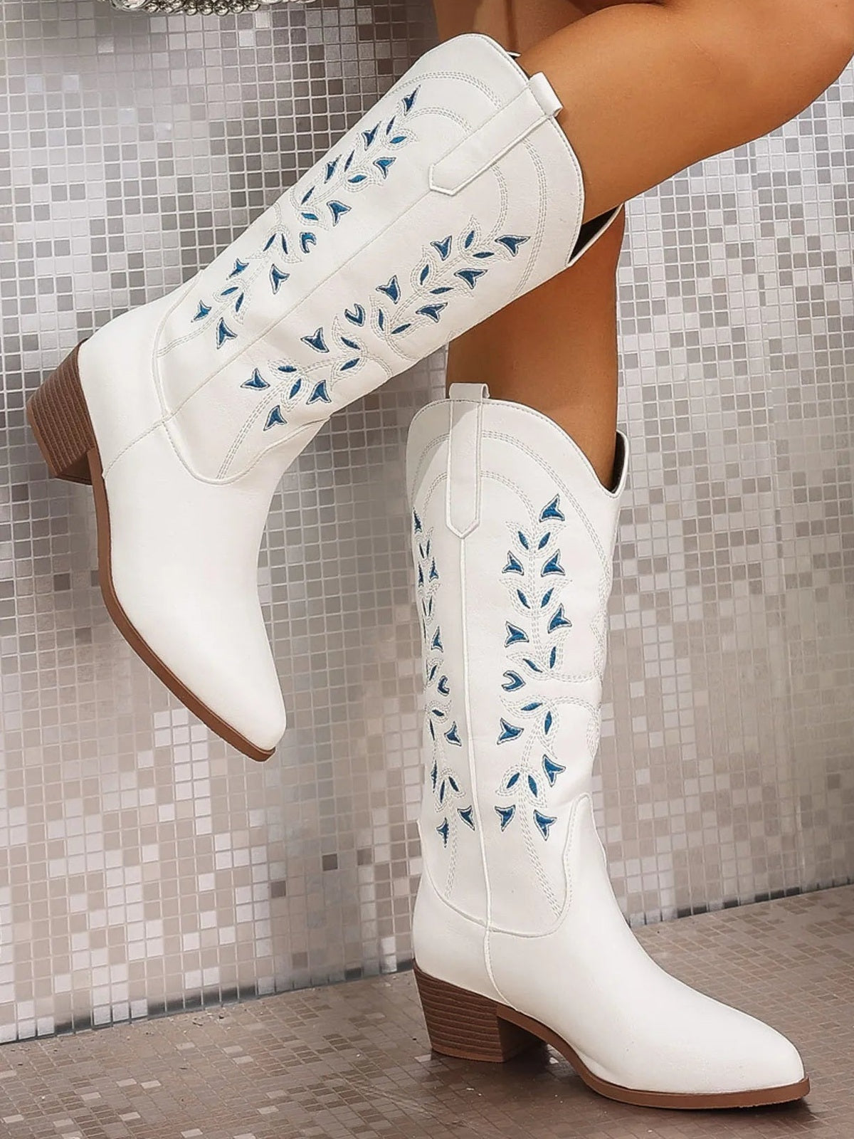 White PU leather point-toe block-heel boots with tall shafts, blue floral stitching, curved panel seams, pull tabs, and stacked mid heels shown on crossed legs.