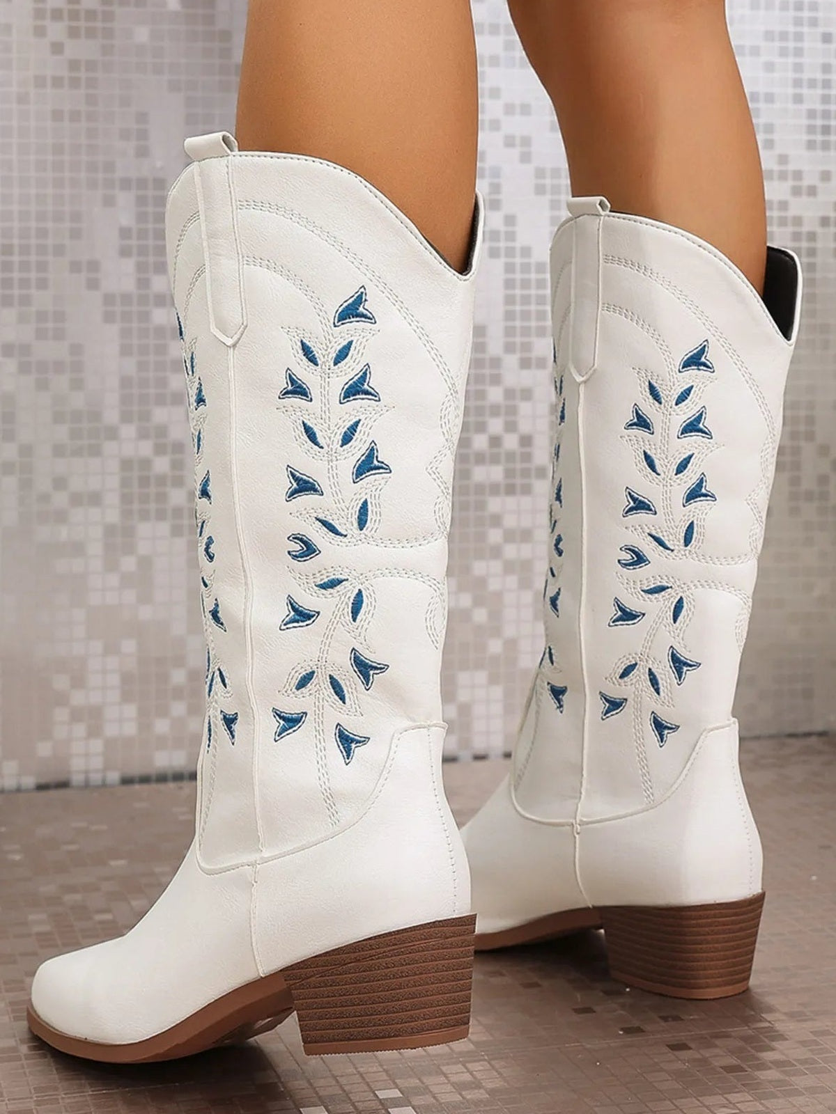A model wears white PU leather mid-calf block-heel boots with blue floral embroidery, stitched panels, pull tabs, and brown rubber soles.