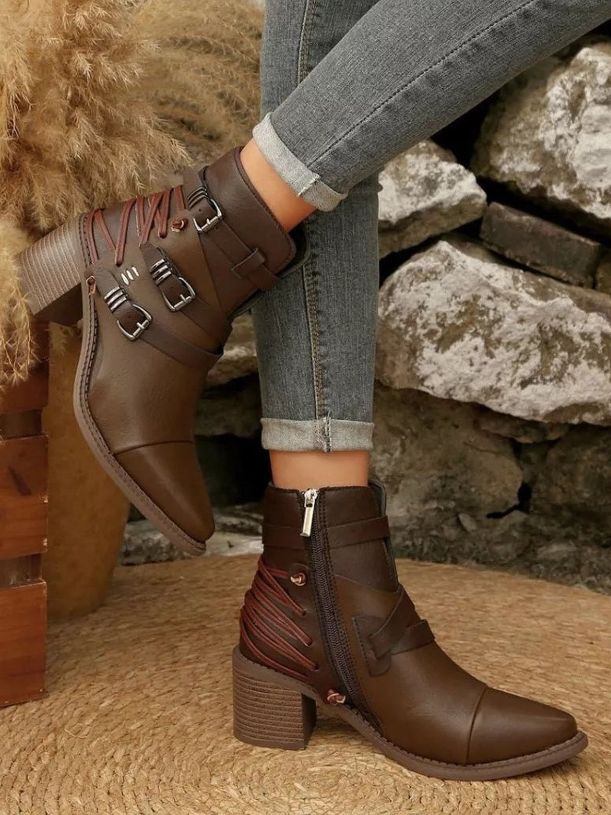 Brown point-toe ankle boots with mid block heels, side zippers, wraparound straps, and multiple metal buckles, modeled with crossed legs.