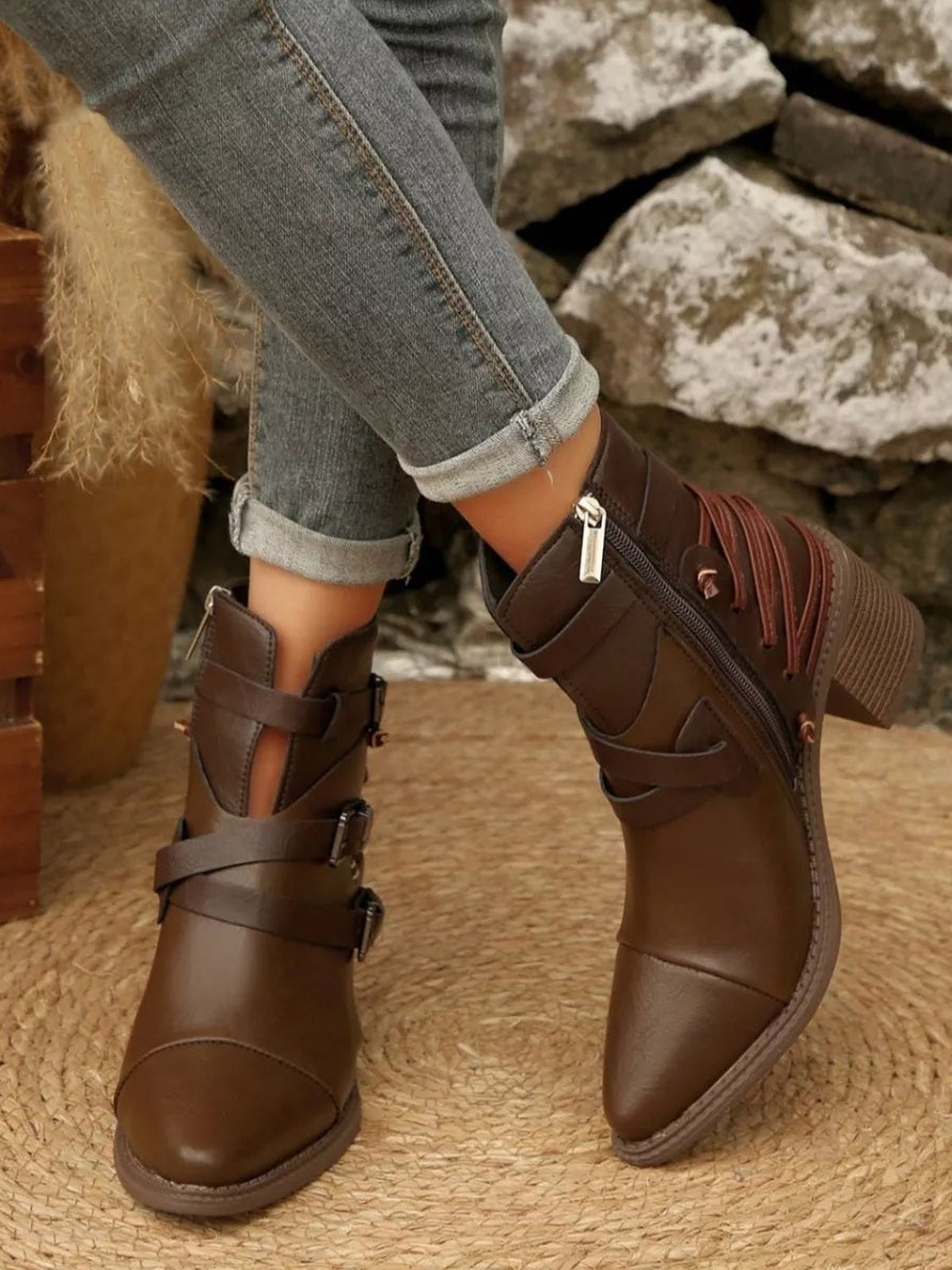 A model wears brown PU leather ankle boots with wraparound buckle straps, side zippers, textured rubber soles, and rolled-up gray jeans.