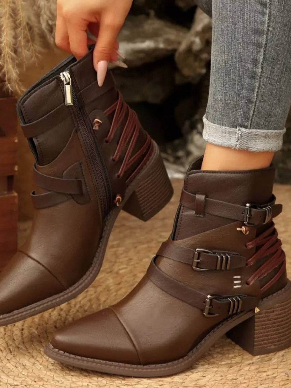 Brown point-toe ankle boots feature multiple buckle straps, side zippers, lace-up accents, and textured mid-height block heels, with one boot held by the model.