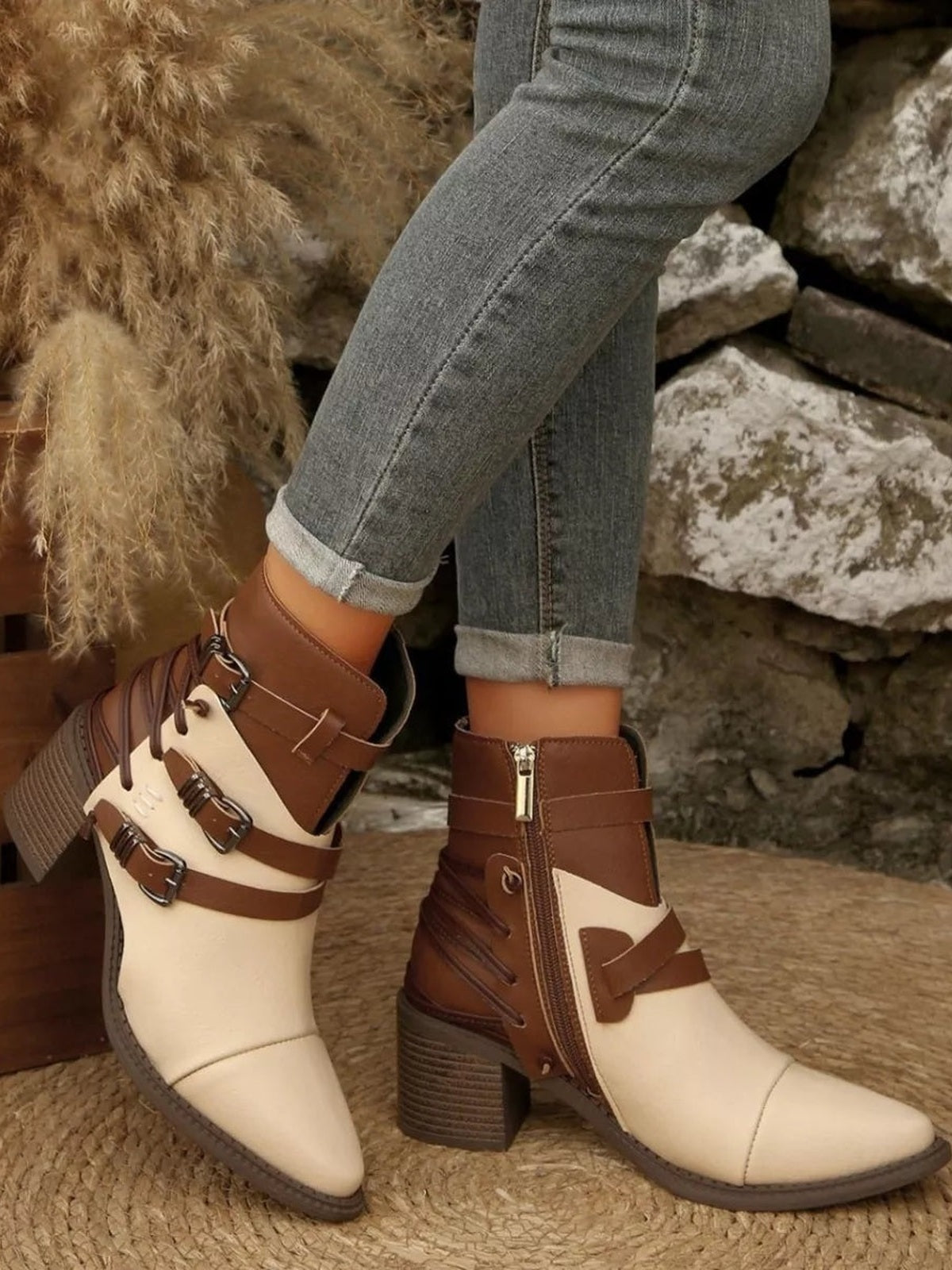 A model crosses her legs to display two-tone cream and brown point-toe ankle boots with buckle straps, side zippers, and sturdy mid-height block heels.