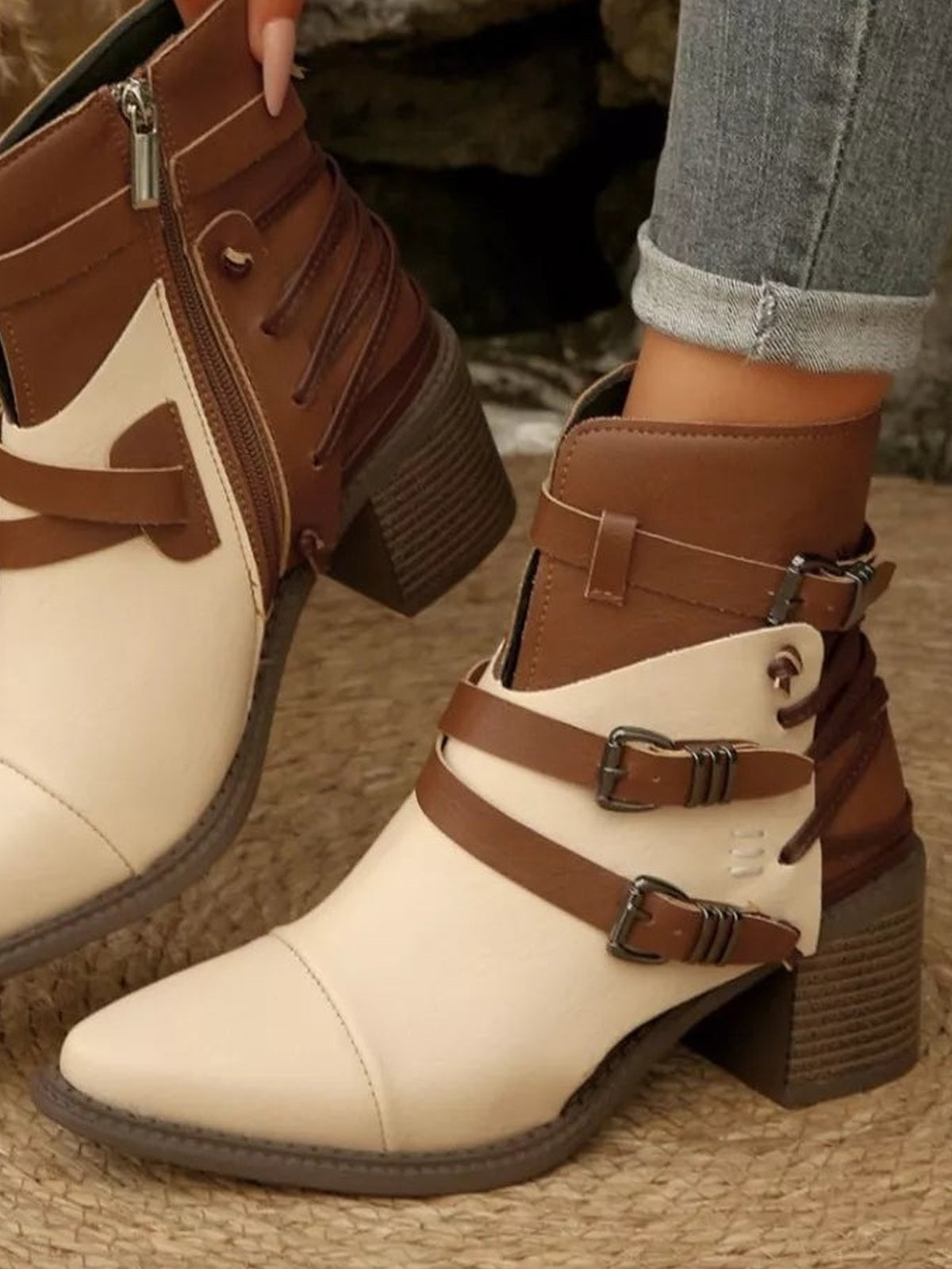 A model wears cream and brown point-toe ankle boots with twin buckle straps, side zippers, contrasting panels, and textured block heels.